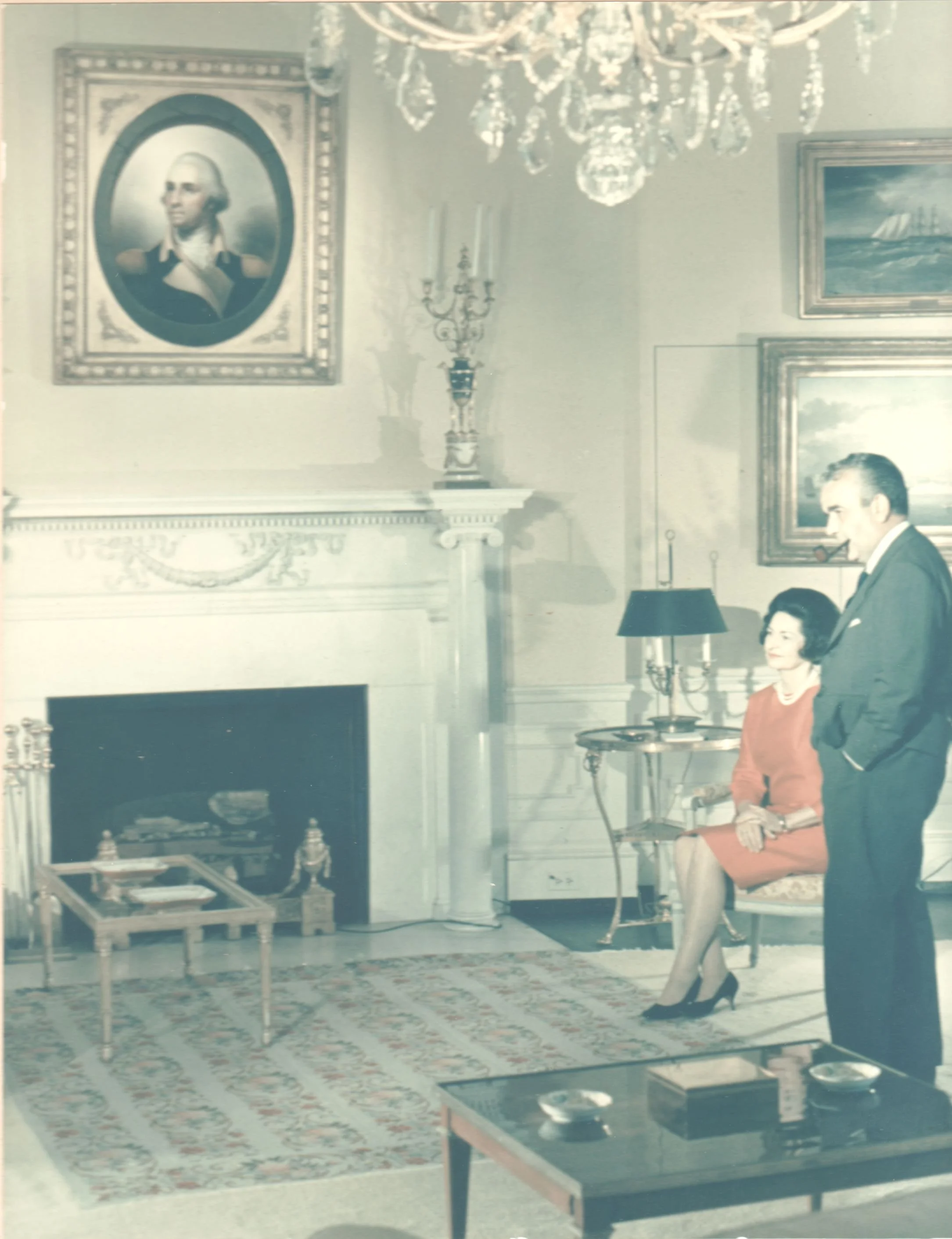 A vintage photograph of a living room featuring a large portrait of George Washington above a fireplace, a chandelier, an ornate rug, and a man and woman dressed in 20th-century attire standing in the room.