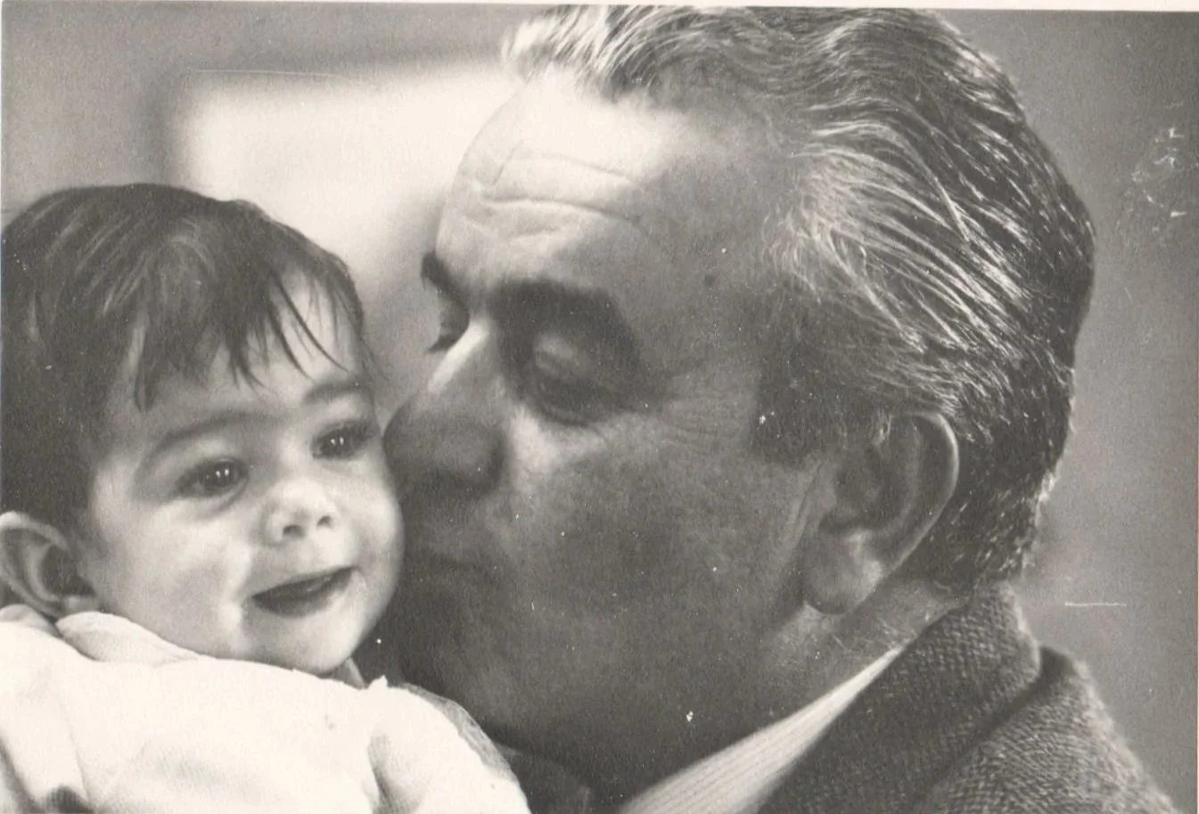 A black and white photograph of a man with slicked-back hair kissing a young boy on the cheek, both smiling.