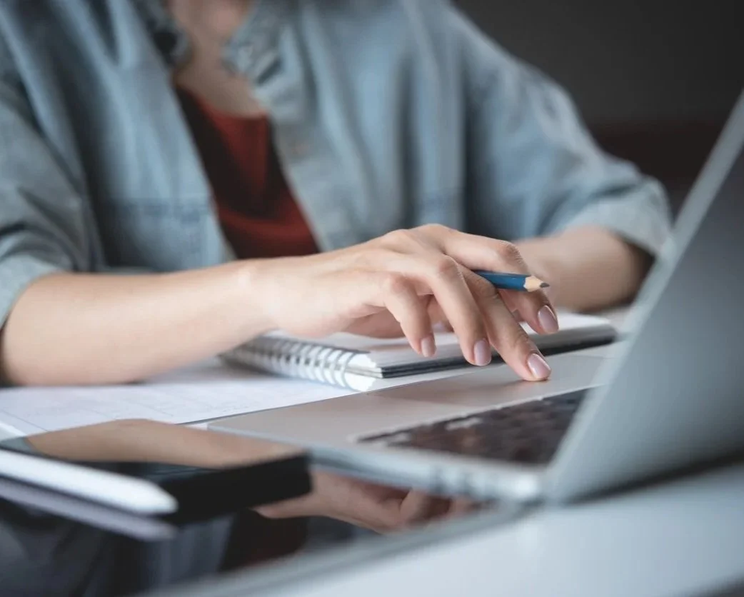 Close-up of a person's hand using a laptop with a notebook, pen, and papers on the desk.