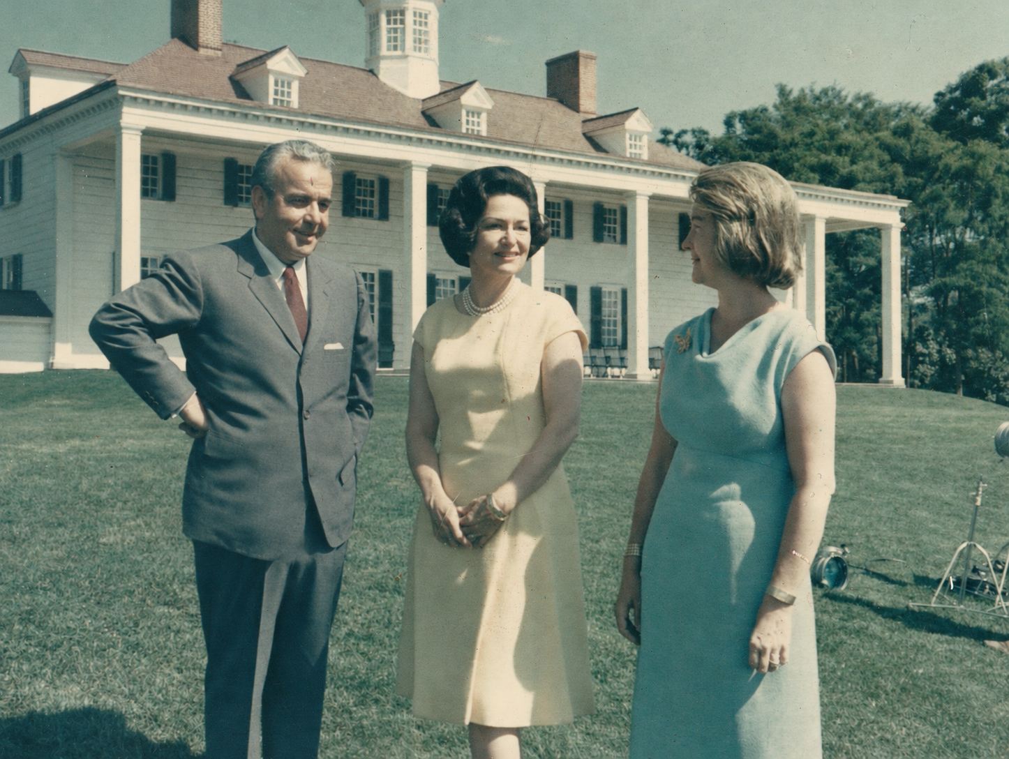 Three people standing on a lawn in front of a large, white, historic house with columns. The man on the left is wearing a gray suit, the woman in the middle is in a yellow dress with pearl necklace, and the woman on the right is in a light blue dress. They appear to be engaged in conversation outdoors on a sunny day.