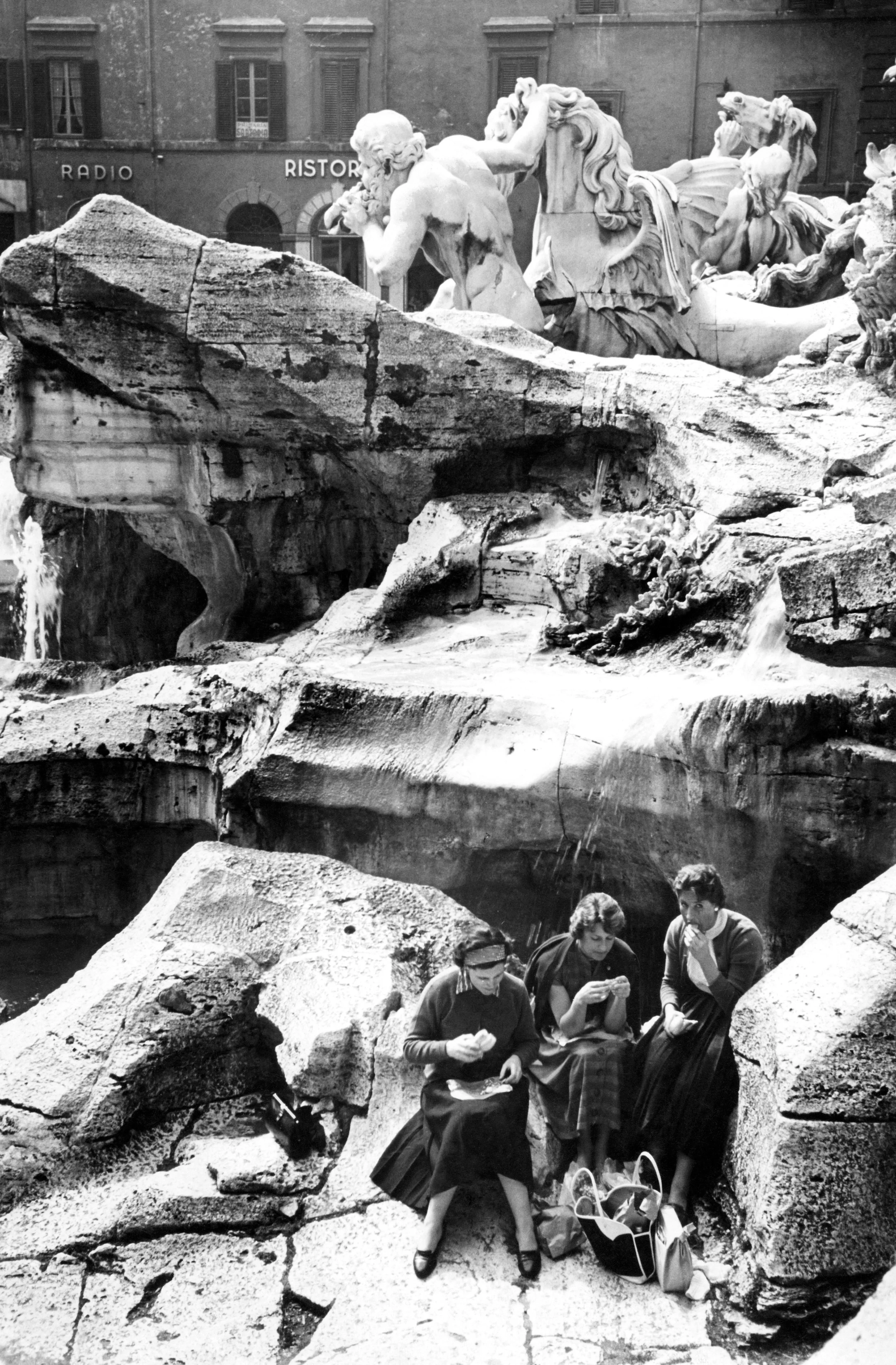 Three women sitting on rocks near a fountain, surrounded by large stone blocks, with backpacks on the ground and water flowing from the fountain. In the background, there is a building with European-style windows and a sign for a radio and restaurant.