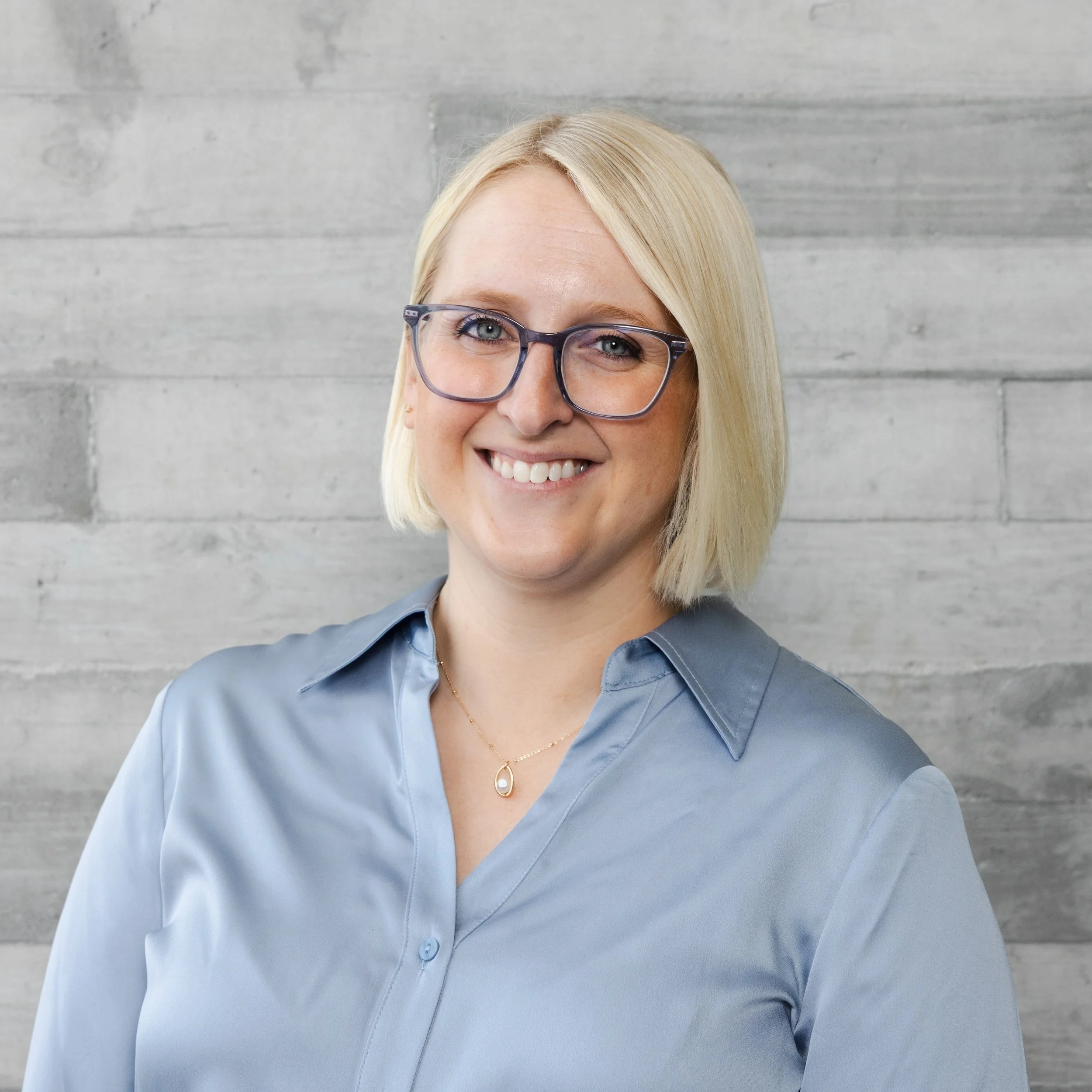 Portrait of a woman with blonde hair, blue eyes, smiling, wearing a blue blouse, standing against a wooden background.