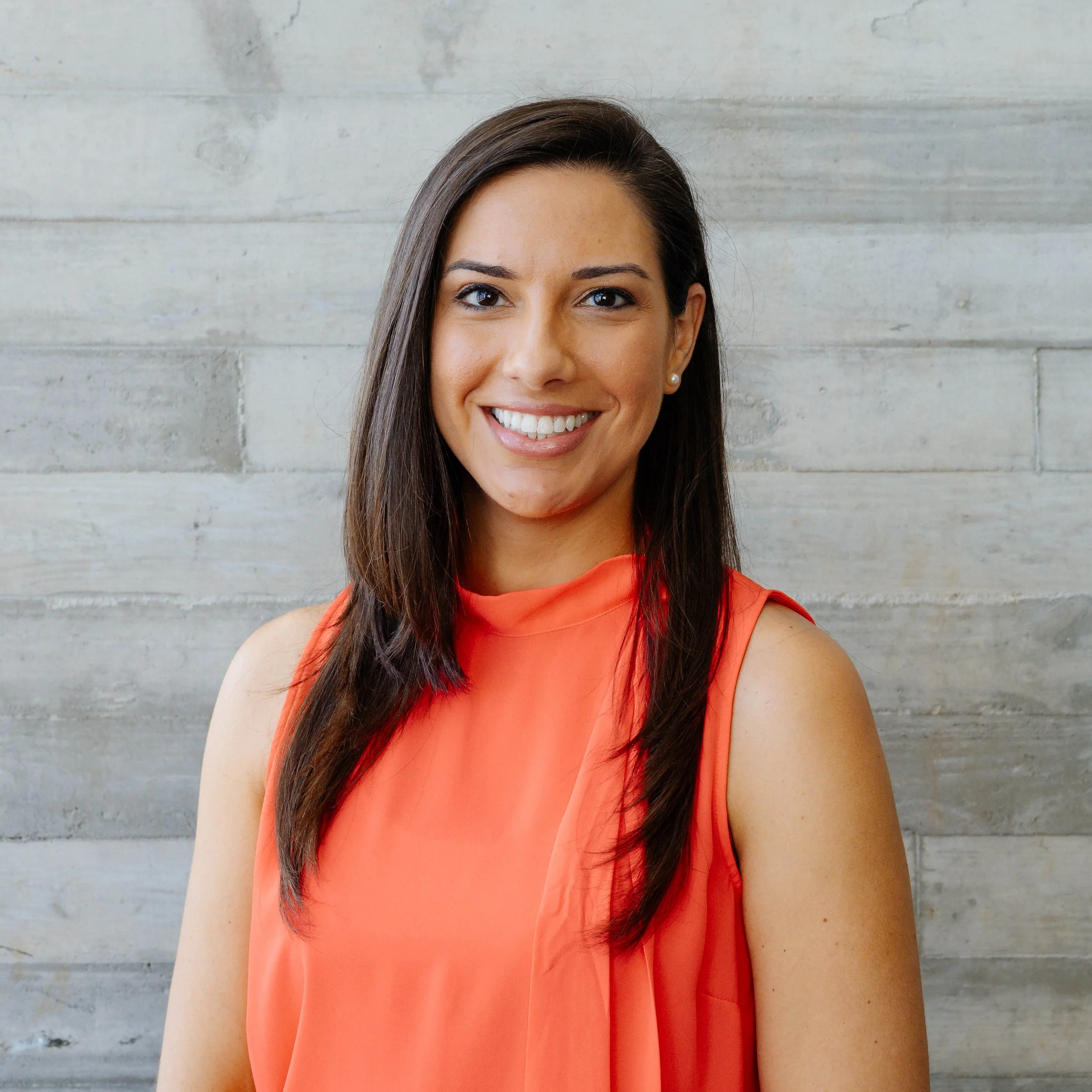 A smiling woman with long dark hair wearing an orange sleeveless top, standing in front of a light-colored wooden wall.