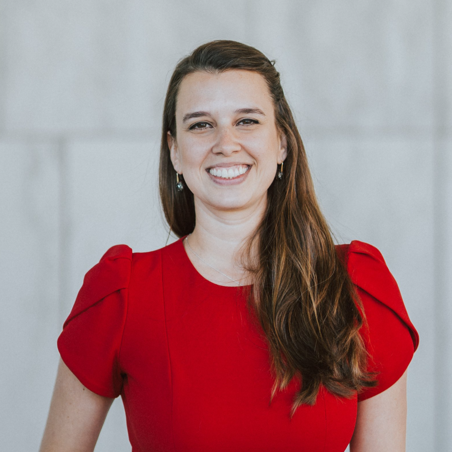 A woman with long brown hair smiling, wearing a red dress and earrings, standing against a light gray wall.