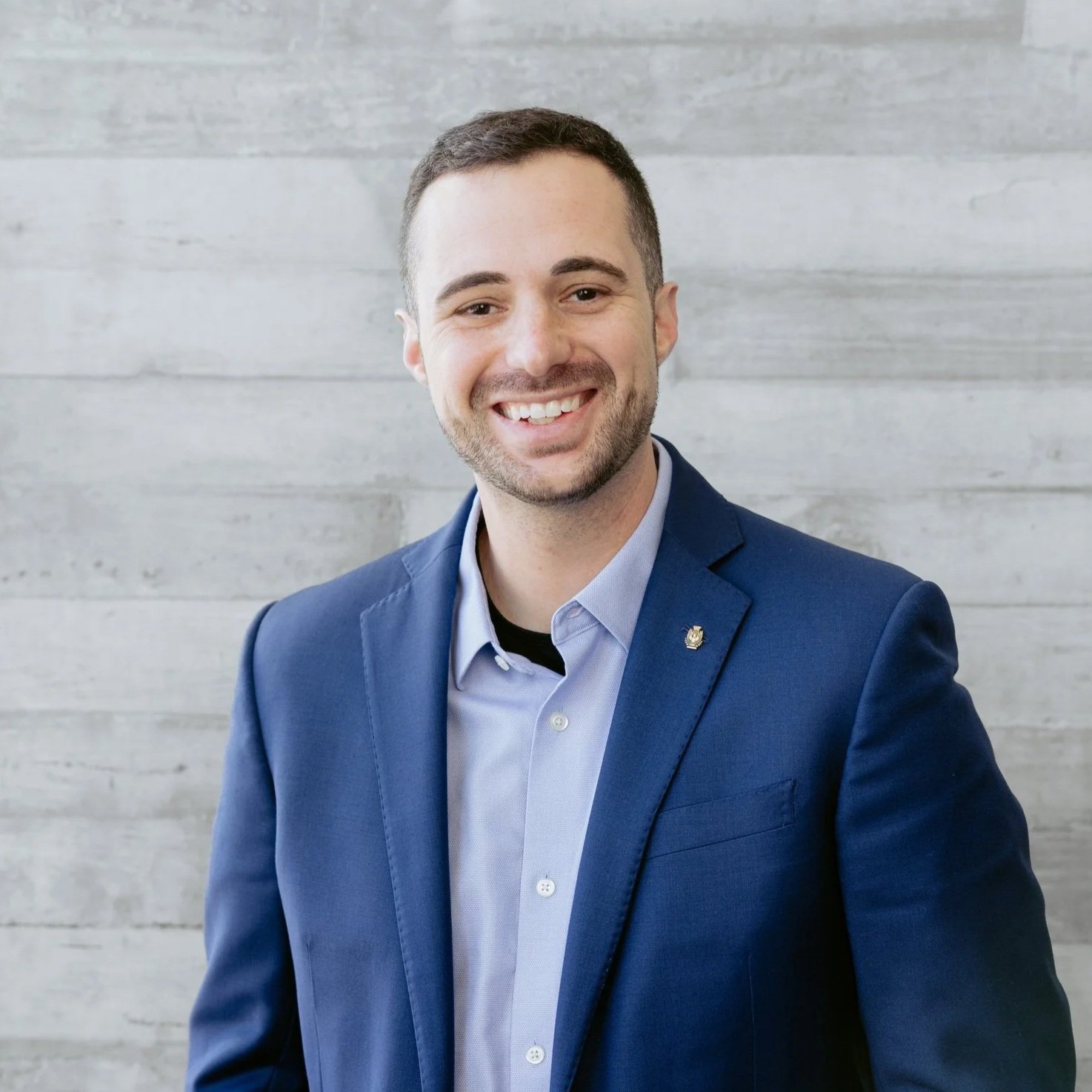 A smiling man in a blue suit and light blue shirt standing against a gray wooden wall.