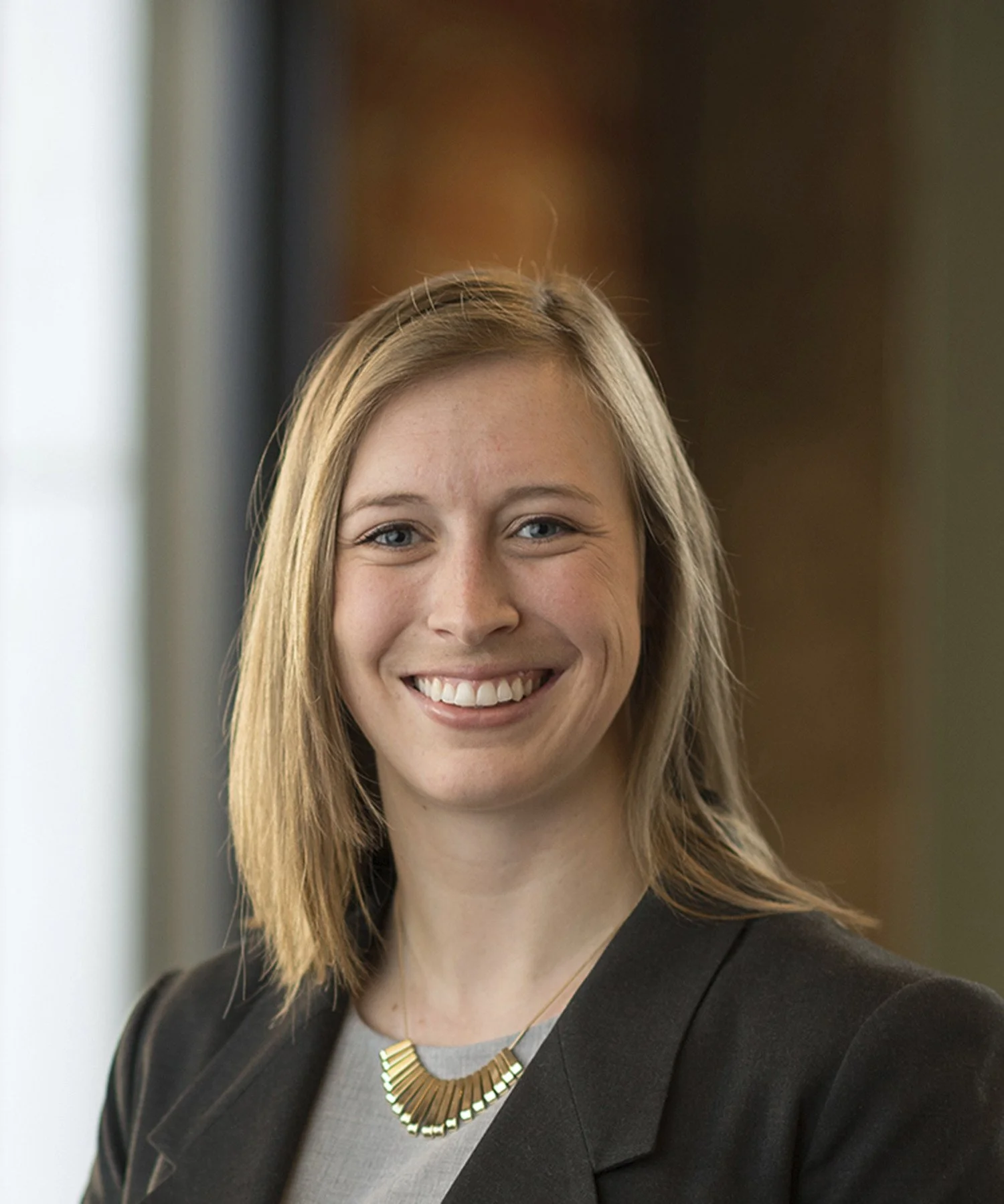 A woman with shoulder-length blonde hair smiling and crossing her arms, wearing a navy blue blouse, in a bright office setting with large windows in the background.