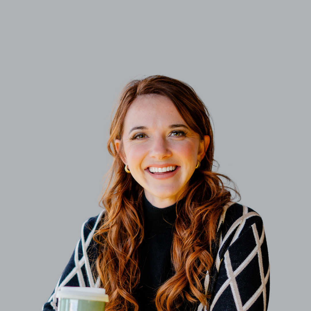A smiling woman with long red hair, wearing a black and white patterned sweater, sitting in a well-lit indoor setting, holding a paper coffee cup.