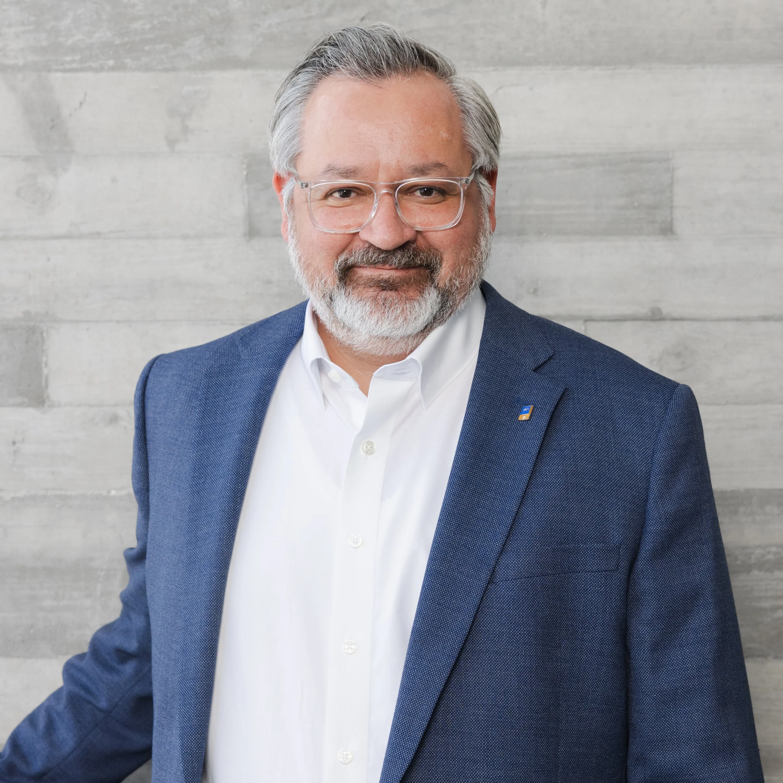 A middle-aged man with glasses, a beard, wearing a white shirt and a blue blazer, standing in front of a textured gray wall.