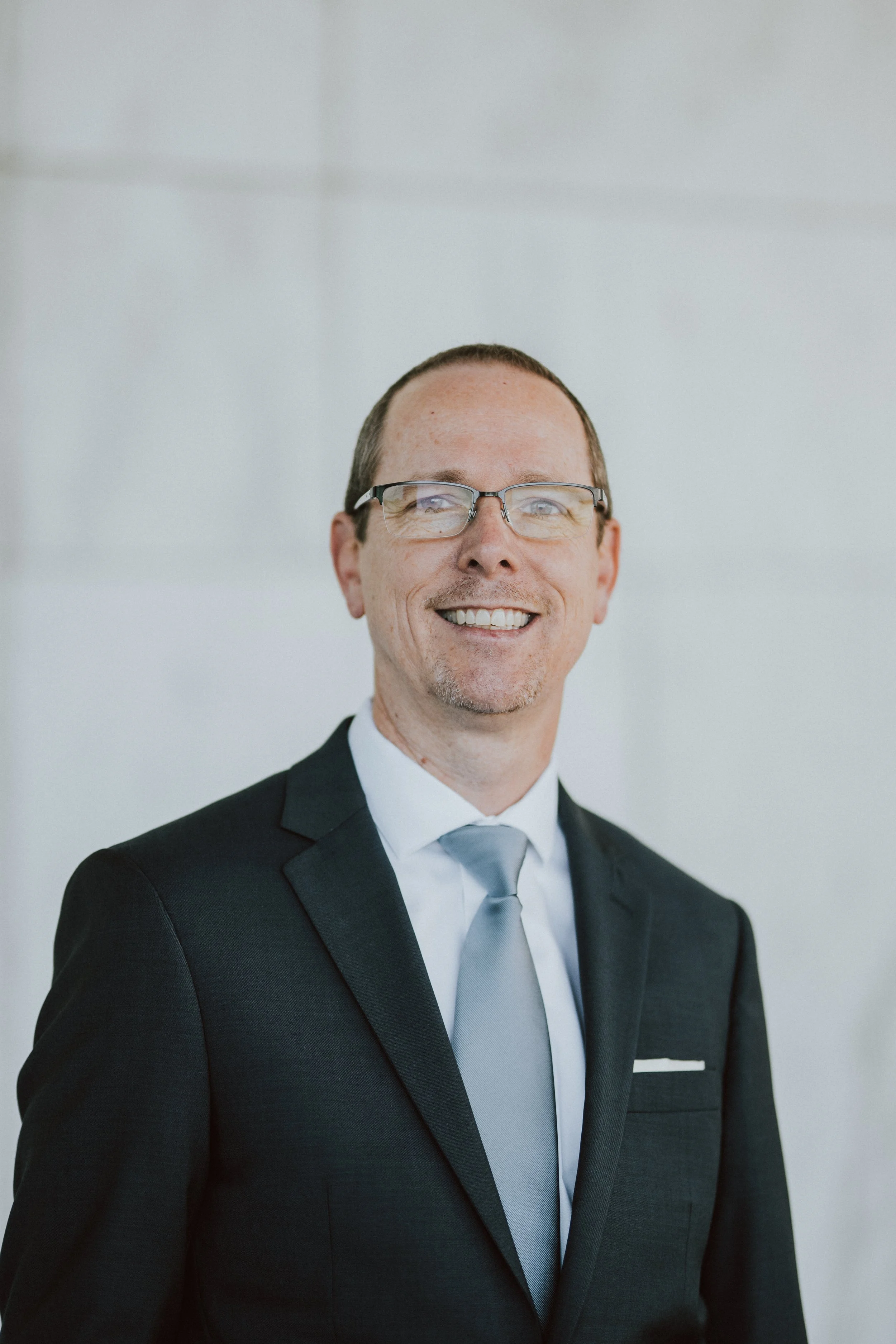 A professional headshot of a middle-aged man with a bald head, light beard, wearing a grey checked suit and a light blue shirt, smiling slightly against a plain light grey background.