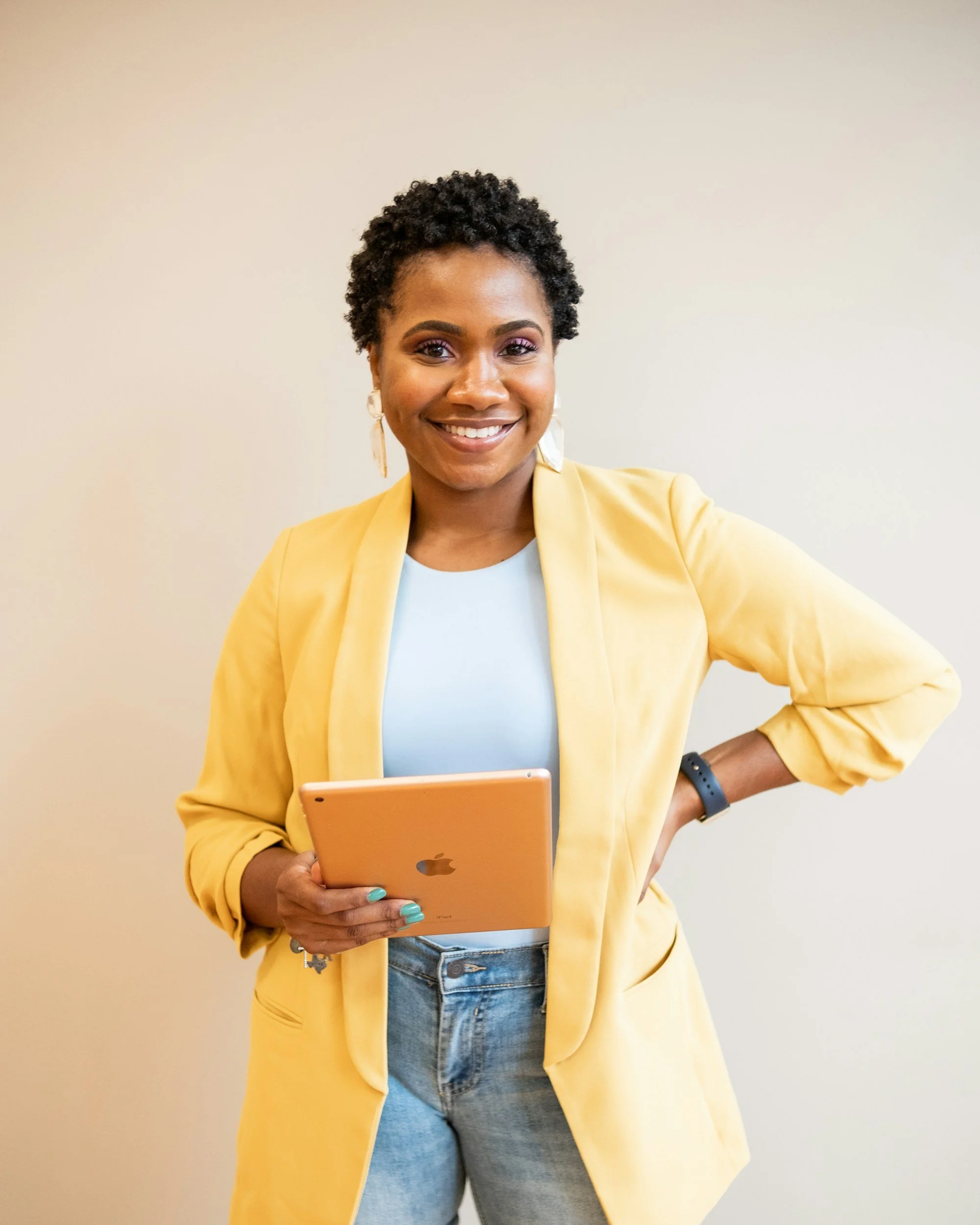 A woman with curly black hair, wearing a yellow blazer, light blue top, and jeans, holding a pink tablet, standing against a plain beige wall.