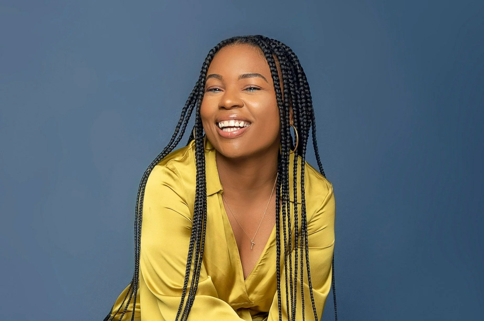A smiling woman with long braided hair wearing a yellow satin blouse and hoop earrings, posed against a blue background.