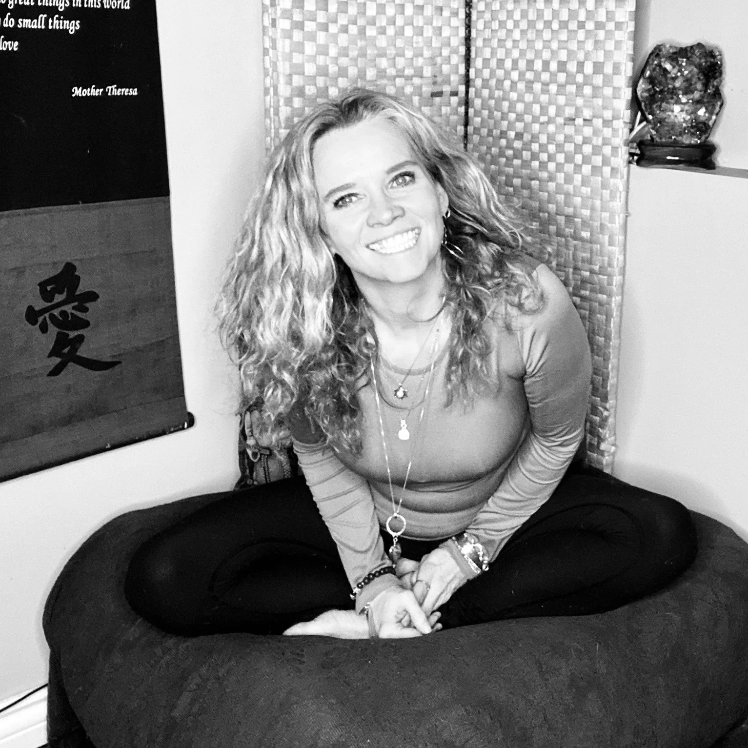 A woman with curly hair smiling, sitting cross-legged on a cushion or bed, wearing multiple necklaces and bracelets, in a room with decorative items and artwork on the walls.