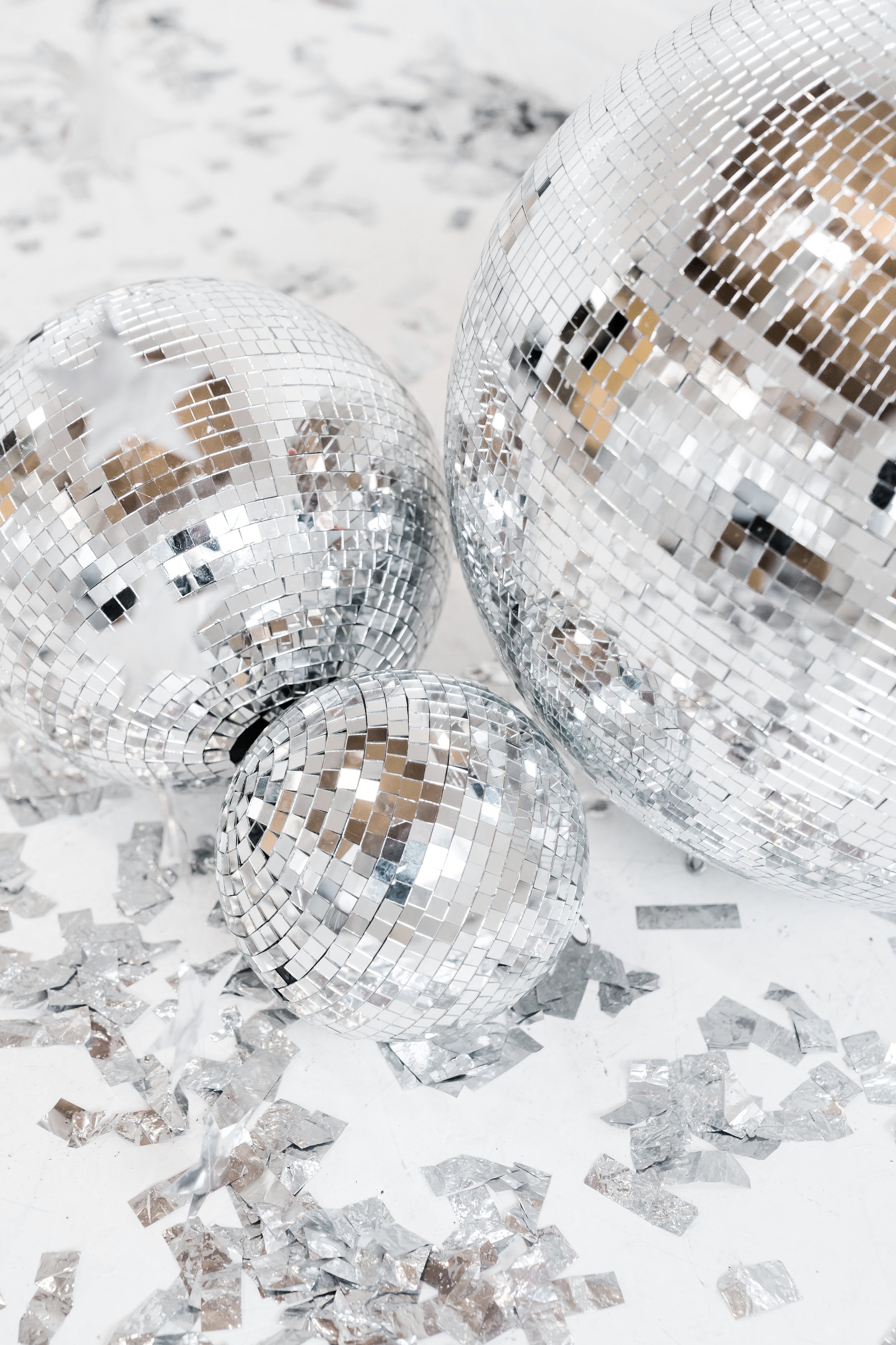 Three disco balls of different sizes with silver reflective surfaces, surrounded by scattered silver confetti on a white surface.