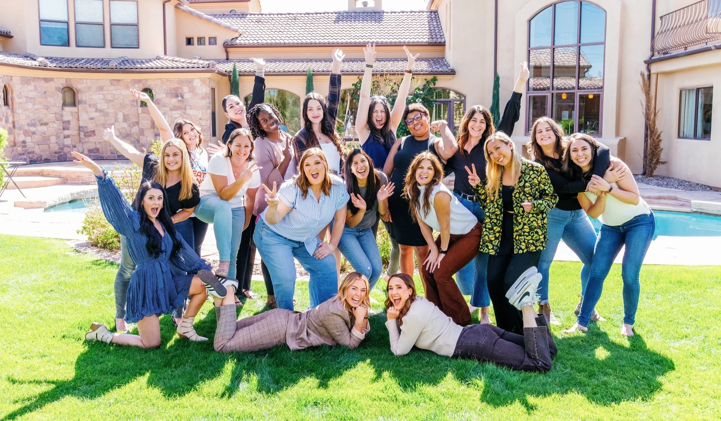 A large group of diverse women posing outdoors in front of a house, smiling and making cheerful gestures on a sunny day.