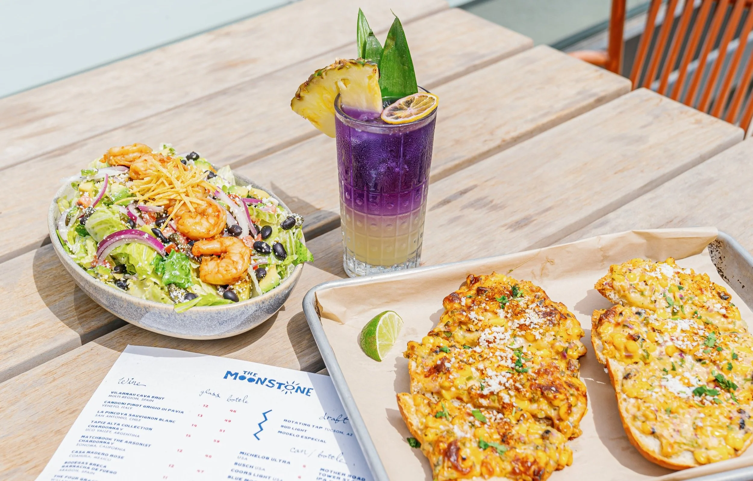 A colorful seafood salad with shrimp, black beans, and vegetables, a layered purple and yellow cocktail with pineapple and lemon garnishes, and two slices of cheesy pizza with corn and herbs on parchment paper, all on a wooden table.
