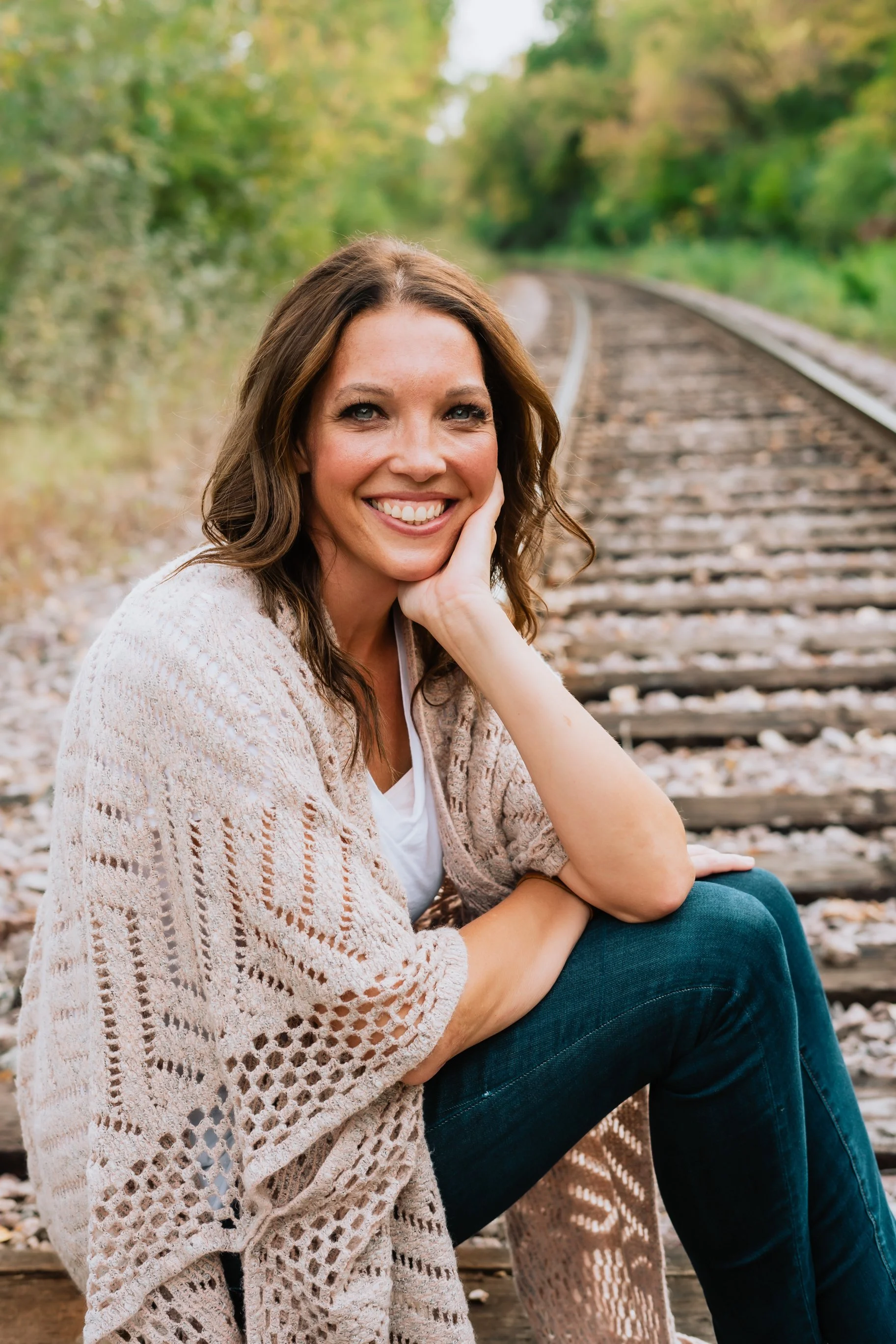 A woman with brown hair, smiling, sitting outdoors on railroad tracks surrounded by trees.