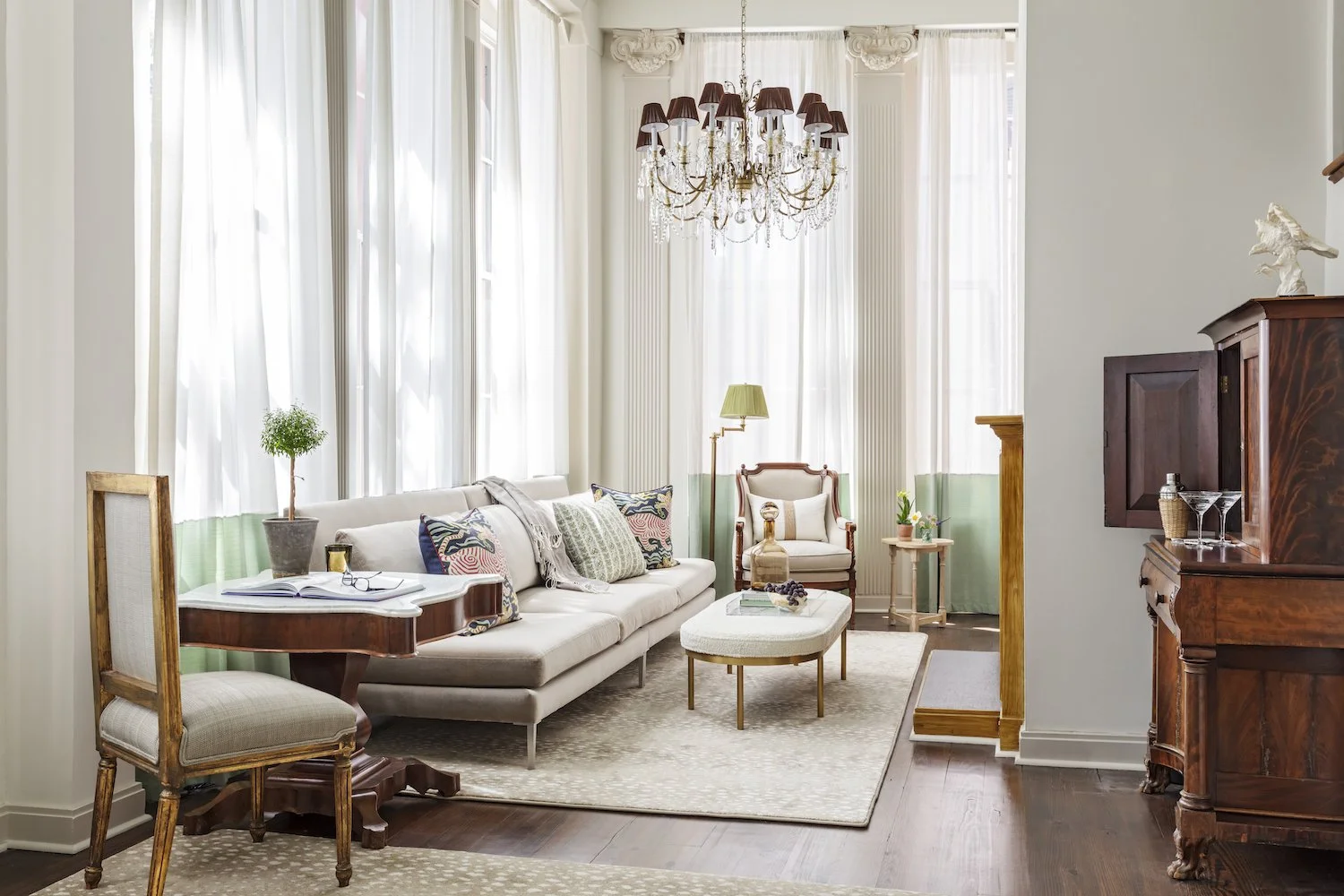 Elegant living room with large windows, white curtains, chandelier, beige sofa with decorative pillows, wooden chairs, side tables, and a patterned rug.