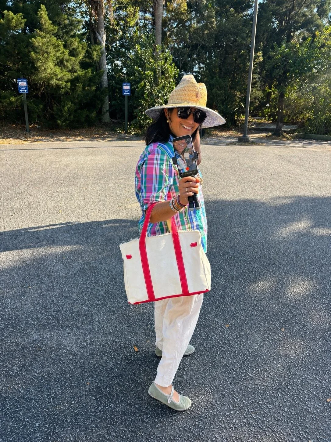 A woman standing outdoors on a paved area, smiling at the camera. She is wearing a large sunhat, dark sunglasses, a colorful plaid shirt, and white pants. She carries a large white tote bag with red straps and trim, and has several bracelets on her wrist. Behind her are green trees and some signs.