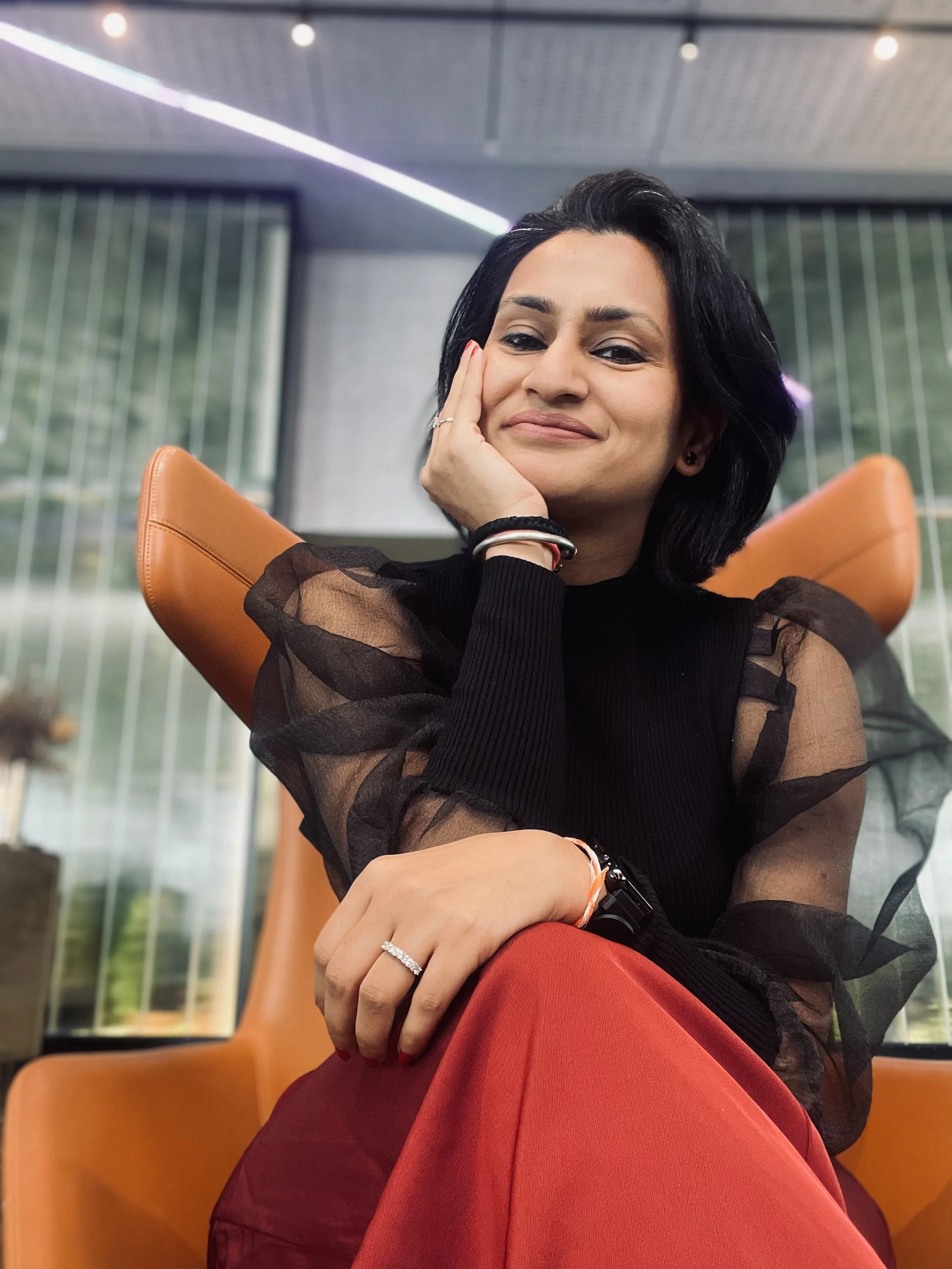 A woman with black hair sits in an orange chair, resting her face on her hand and smiling slightly. She is wearing a black sheer top with puffed sleeves and a ring on her finger. The background shows large glass windows with a modern interior.