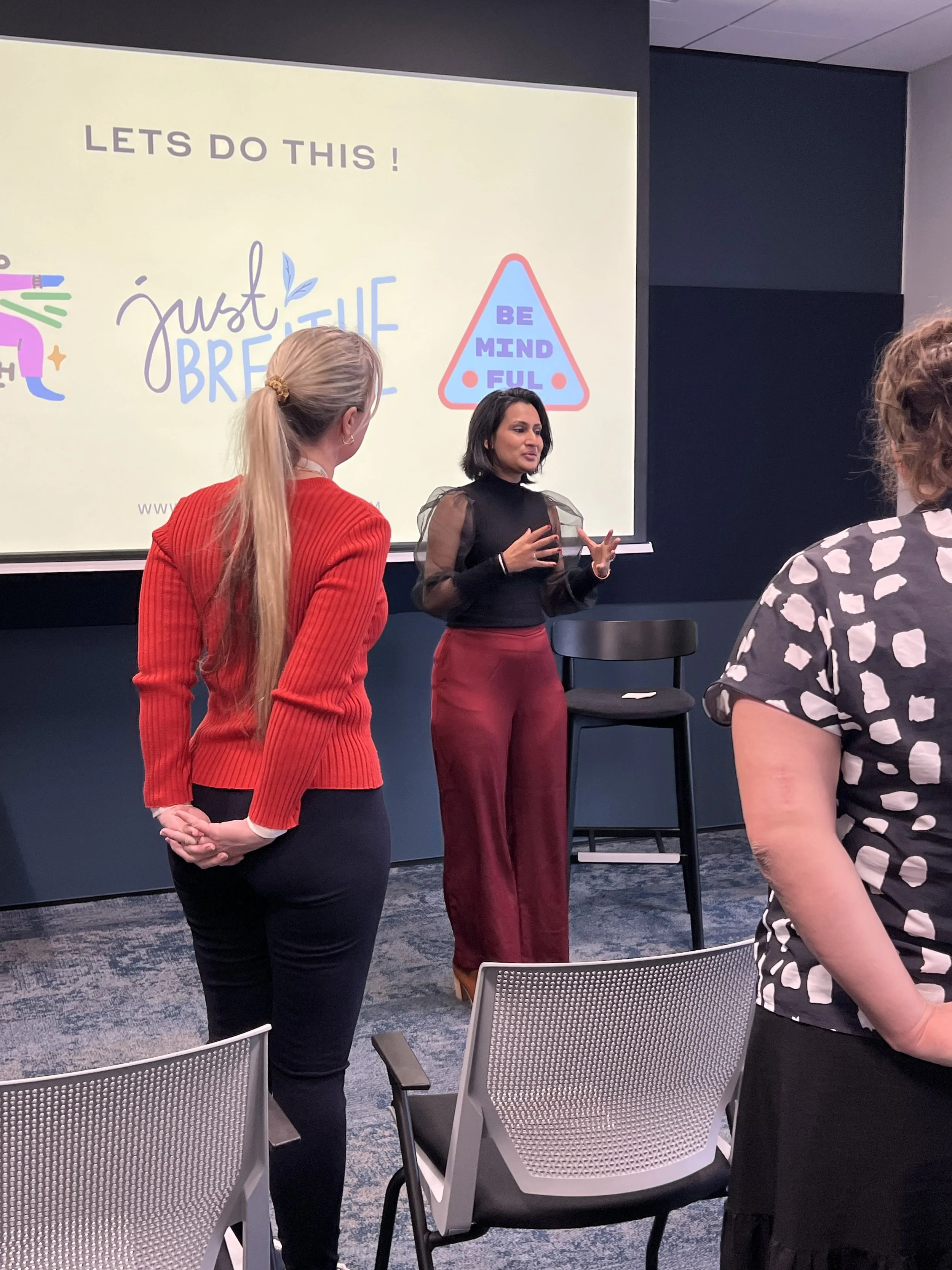 A woman speaking on stage in front of a large screen with a presentation slide that says 'LET'S DO THIS! Just be yourself.' and 'BE MINDFUL'. Two women are listening to the speaker, with chairs and a dark wall in the background.