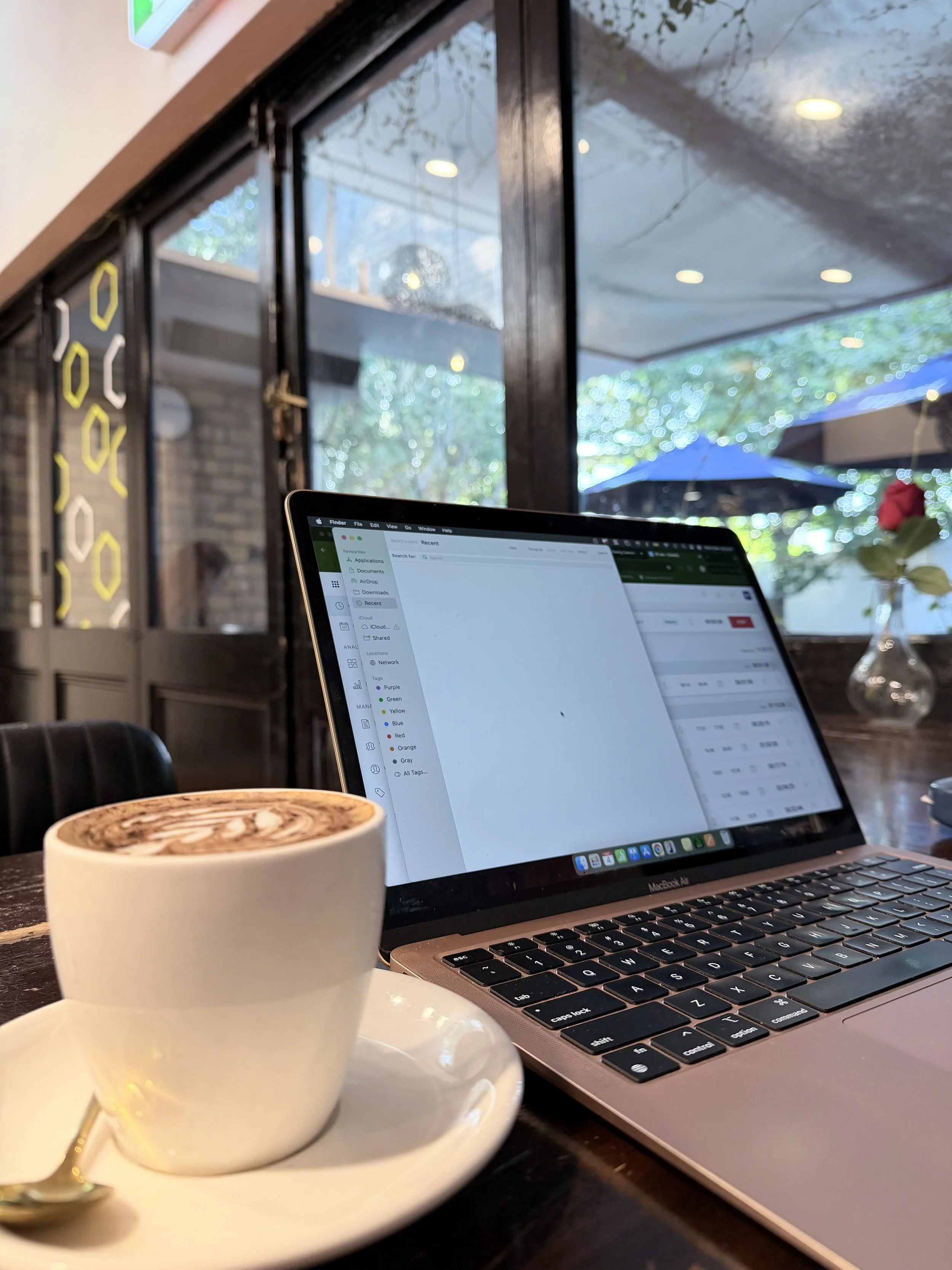 A latte in a white cup on a saucer with a spoon, and an open MacBook Air laptop displaying a spreadsheet, set on a wooden table inside a cafe with large windows, outdoor patio, and trees visible outside.