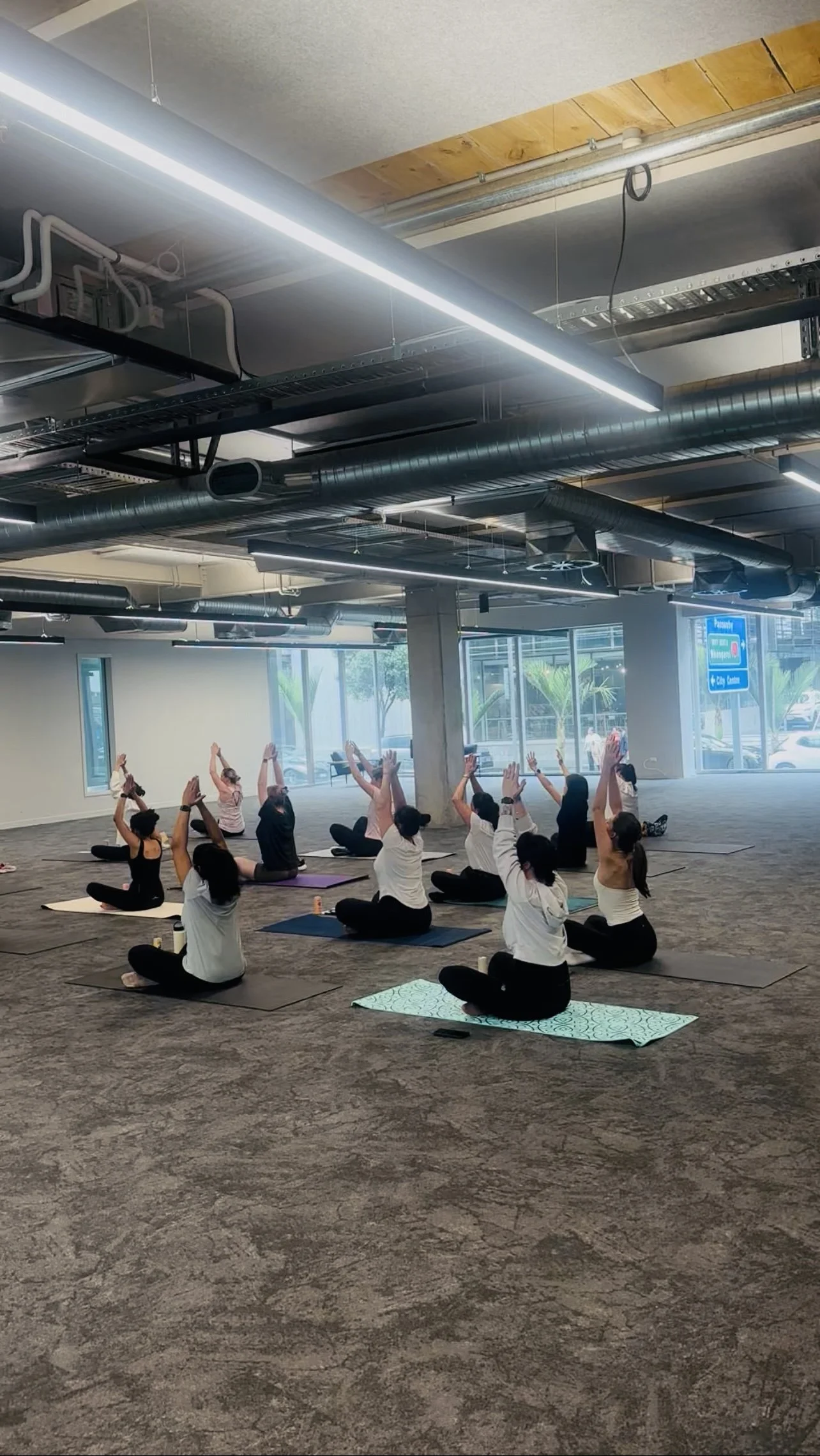 Group of people participating in a seated yoga class in a spacious, modern gym with large windows and industrial ceiling.
