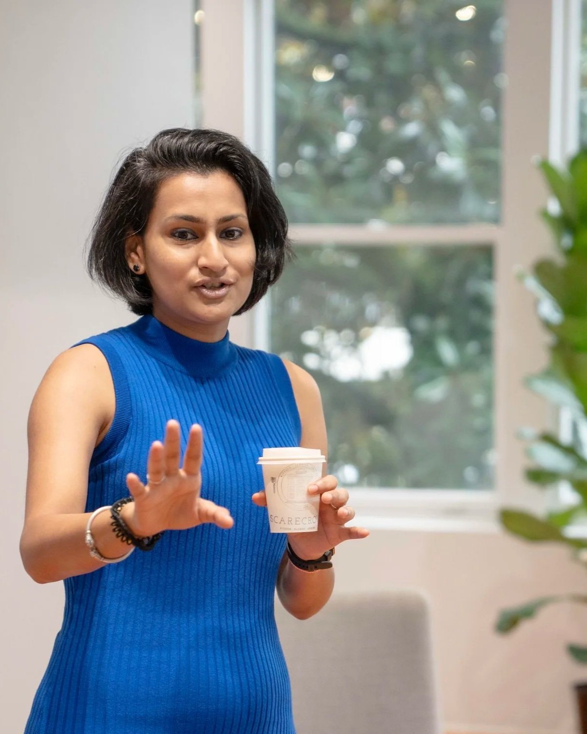 A woman in a sleeveless blue dress holding a white coffee cup and gesturing with her hand indoors near a window with greenery outside.