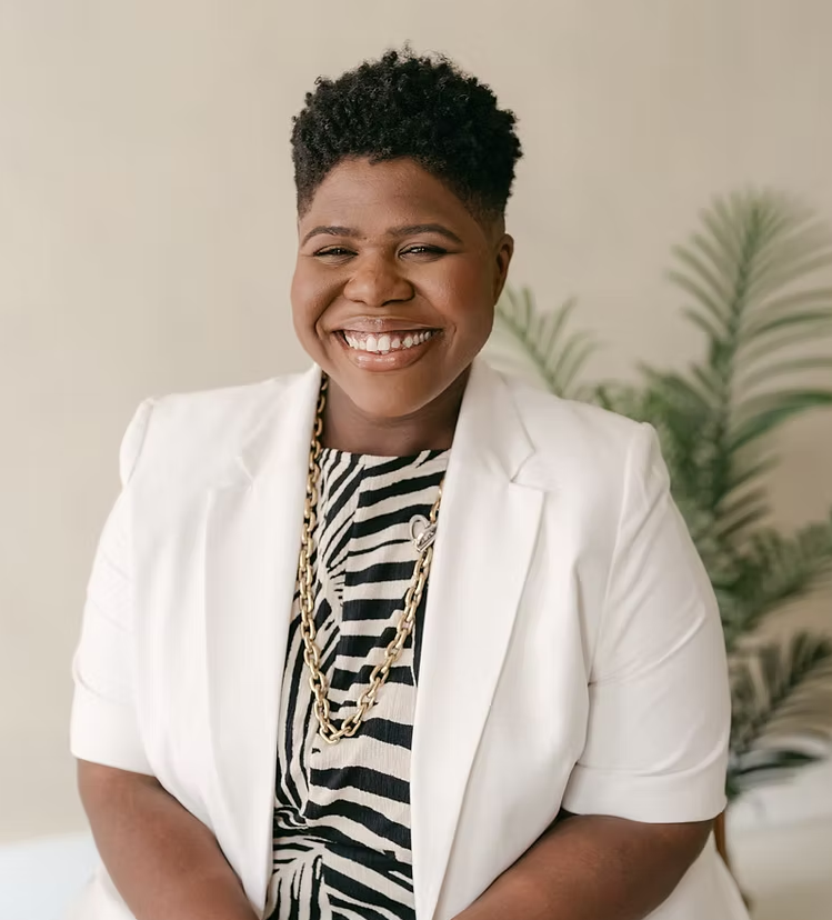 Smiling Black woman with short curly hair, wearing a white blazer, a black and white striped top, and a gold chain necklace, sitting in front of a light-colored wall with a green plant in the background.