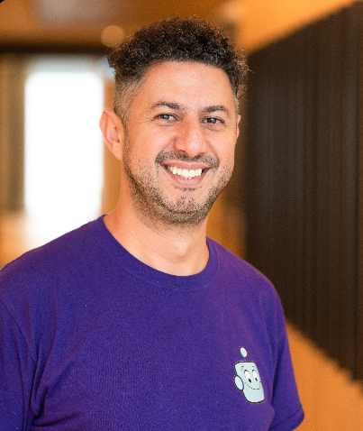 Smiling man with curly hair wearing a purple T-shirt with a robot logo, indoors with warm lighting.