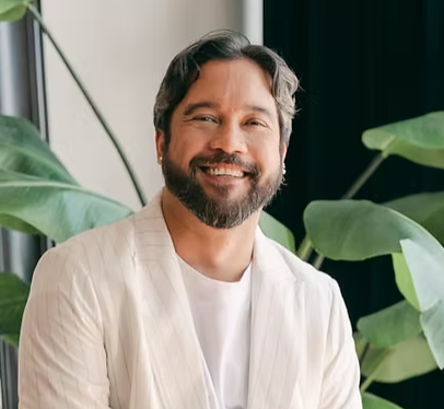 A smiling man with dark hair and a beard, wearing a white blazer, standing indoors near large green plants.