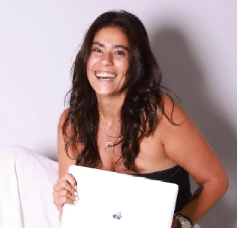 A woman with long dark curly hair, smiling, holding a closed white laptop, sitting on a white bed against a light gray wall.