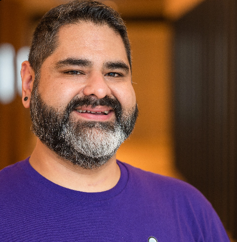Close-up of a smiling man with dark hair, beard, wearing a purple shirt, indoors with warm lighting.