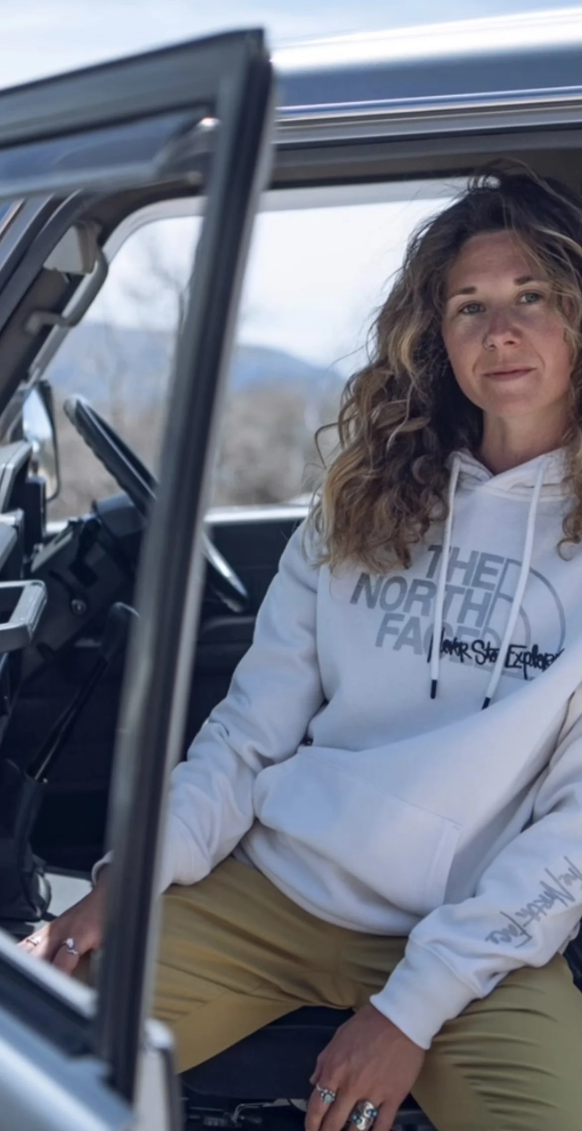 A woman with curly hair wearing a white North Face hoodie sitting in a vehicle with the driver's side door open, with a natural outdoor background.