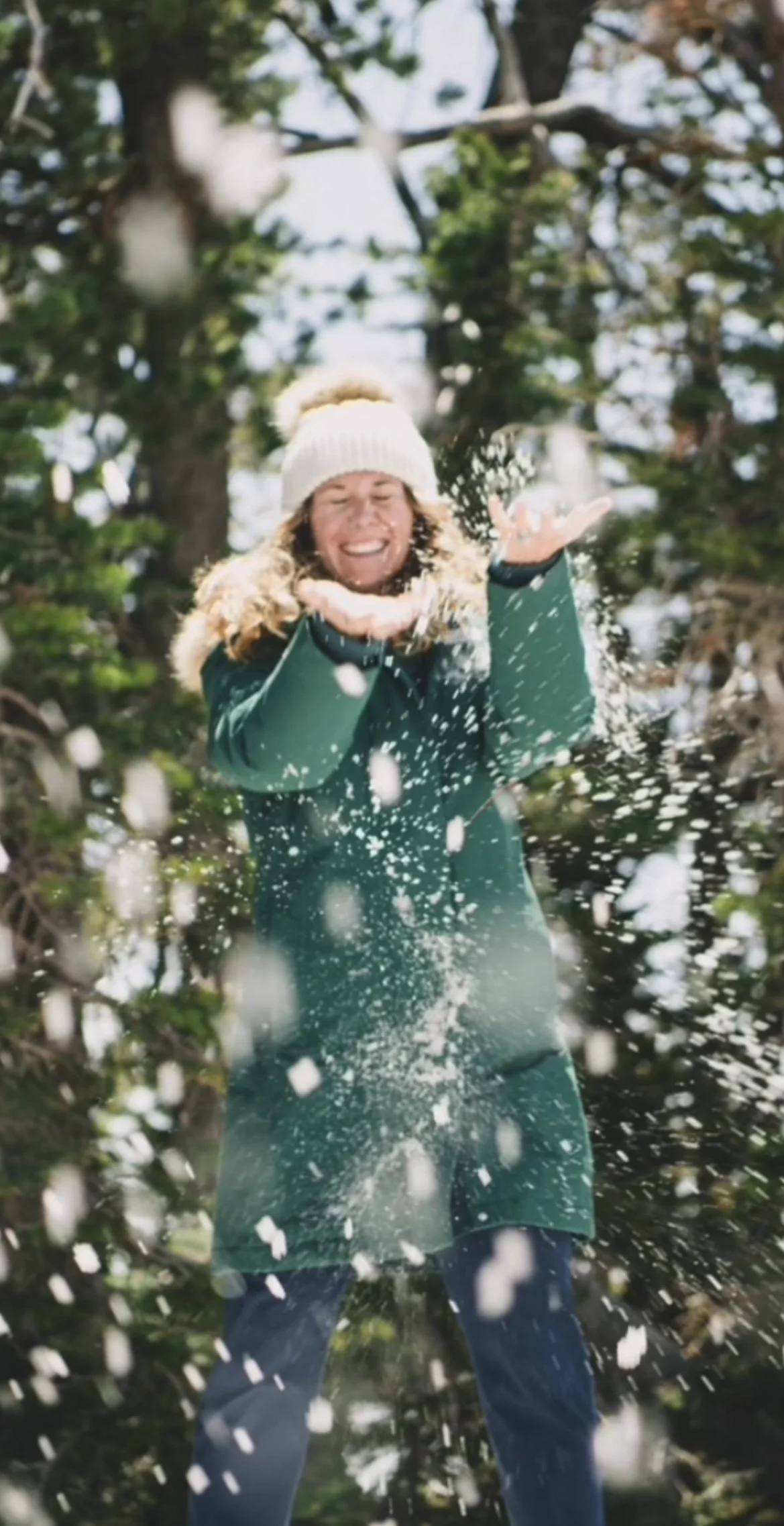 A woman in a white knit hat and green winter coat playing in the snow outdoors, smiling and throwing snowflakes.