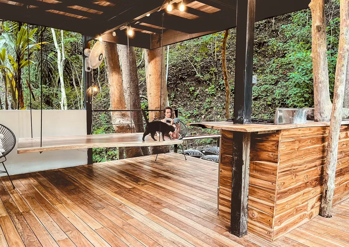 A person lying on a wooden deck with arms outstretched, surrounded by lush green plants, with a black outdoor lounge area with pillows nearby.
