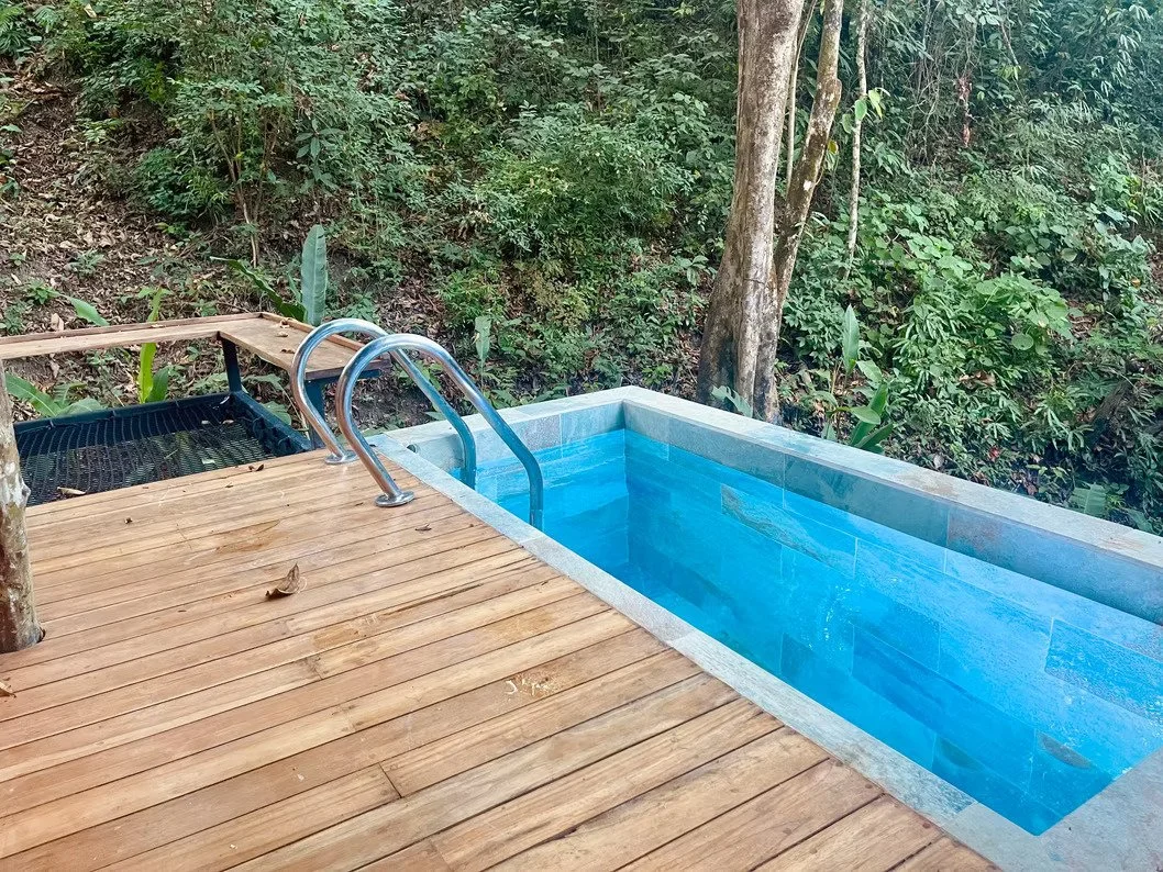 Woman relaxing in a small outdoor pool with a waterfall feature, surrounded by a potted plant and gravel
