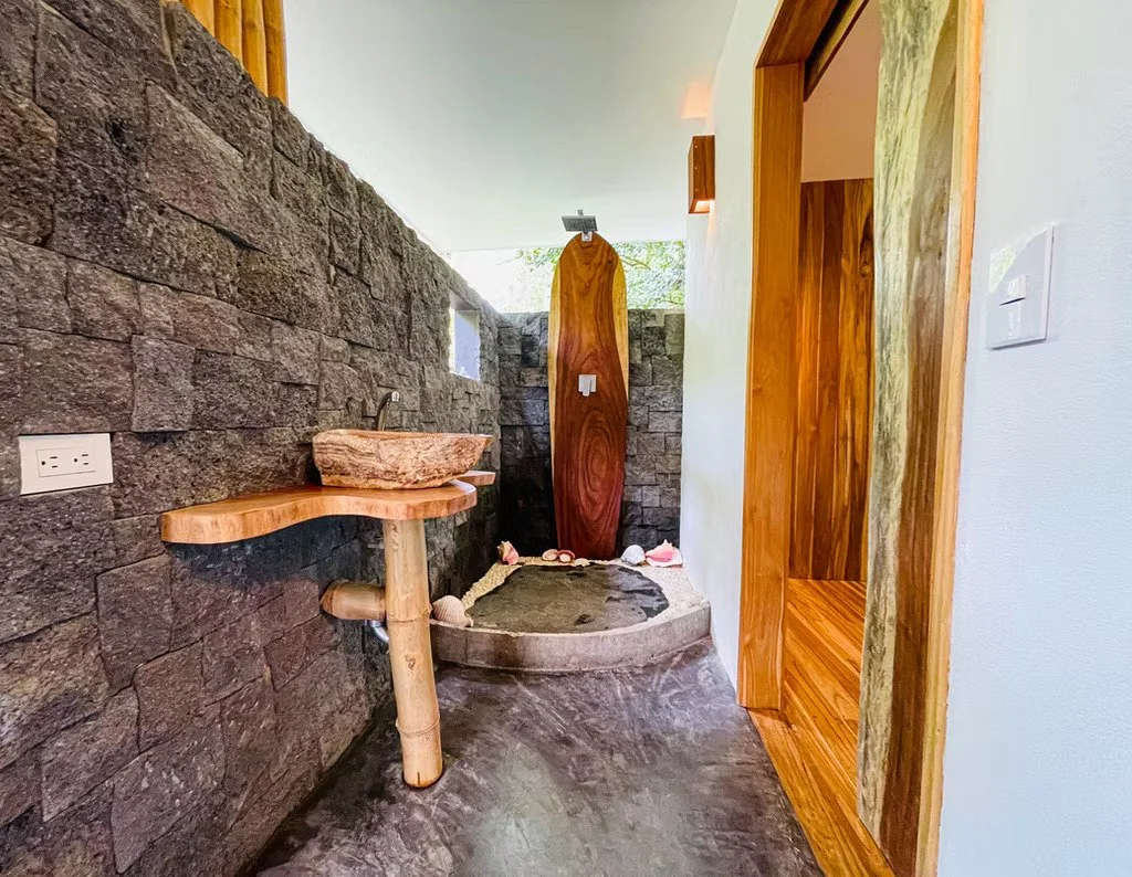 A modern bathroom featuring a white bathtub next to a glass window with plants outside, a small wooden shelf with a faucet, and a wooden door with a curtain.