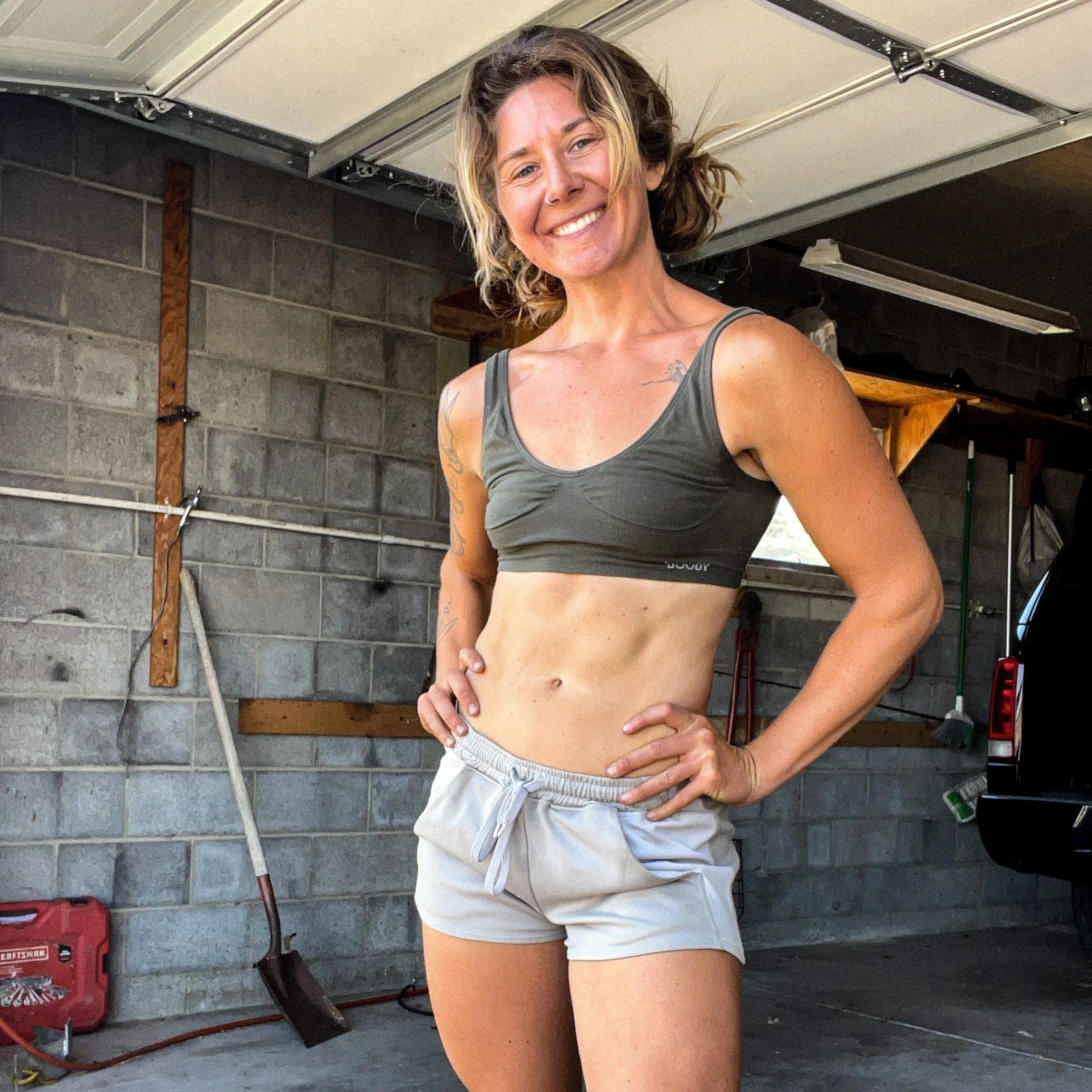 A smiling woman with short, wavy blonde hair, wearing a gray sports bra and light gray shorts, is standing in a garage with her hands on her hips.