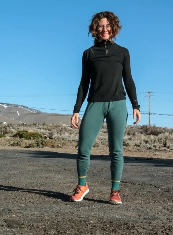 A woman with curly hair and glasses standing outdoors on a dirt road in athletic clothing, smiling, with mountains and power lines in the background under a clear blue sky.