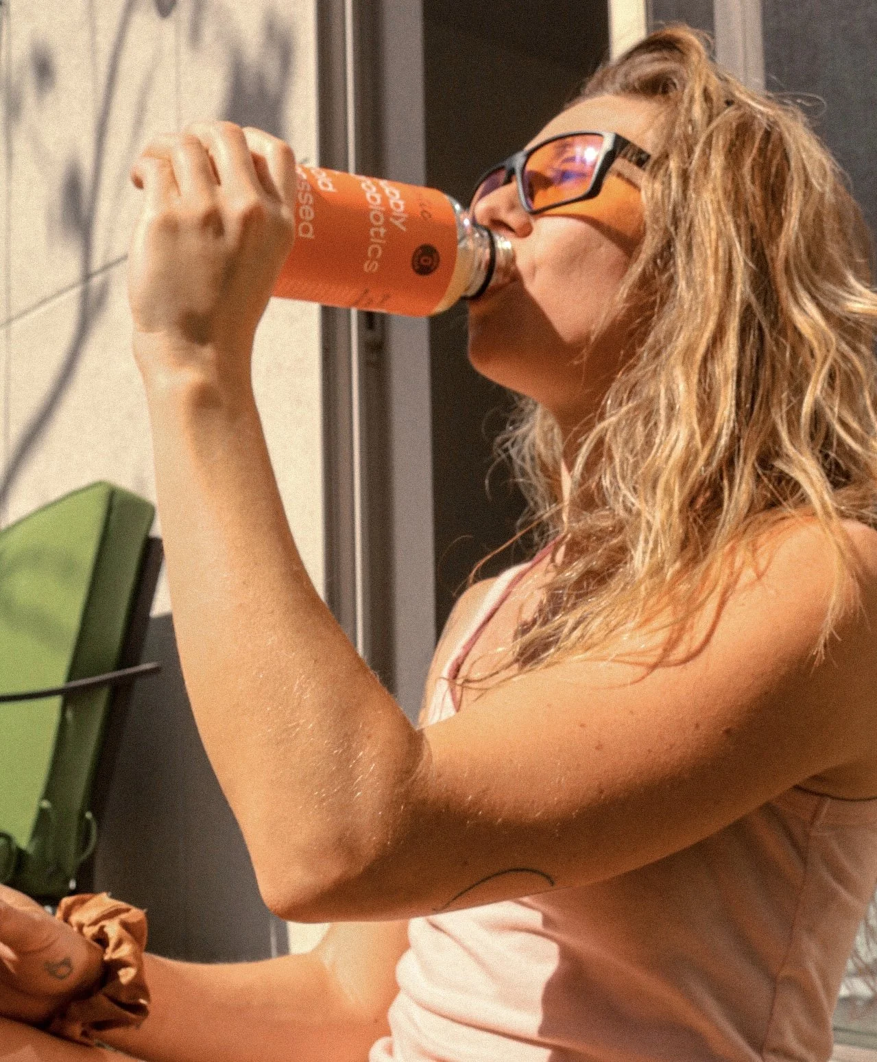 A woman with wavy hair wearing sunglasses drinks from a plastic bottle of sports drink outdoors, sitting on a ledge or bench.