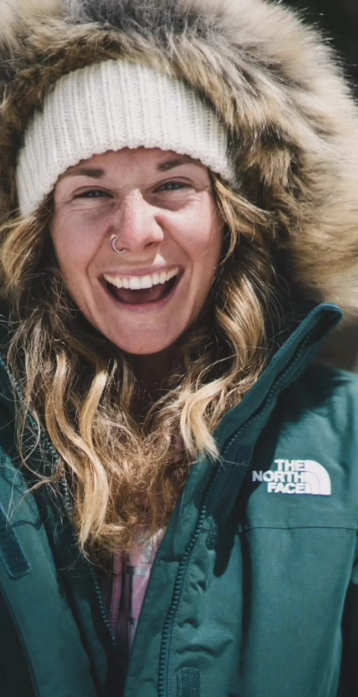 Smiling woman with curly hair wearing a North Face jacket, a white knit beanie, and a fur-lined hood in a winter outdoor setting.