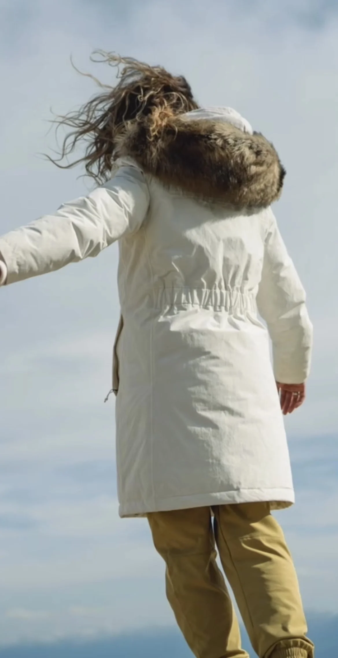 Person in a white winter coat with a fur-lined hood, standing outdoors on a cloudy day with a body of water in the background.