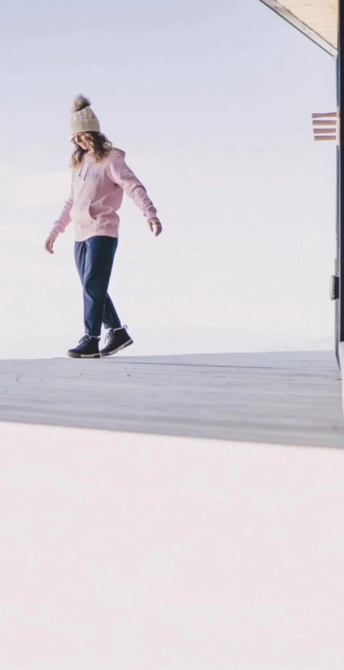 A woman in pink jacket, blue pants, and black boots standing outdoors on a wooden floor, with a ski slope or snowy area in the background, smiling and looking down.