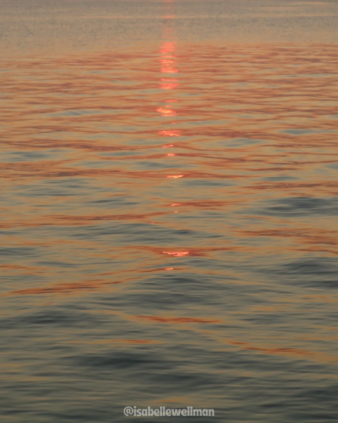 A photograph of a calm body of water at sunset, with the sun’s reflection creating a red streak across the surface.