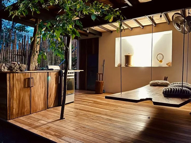 Outdoor patio with wooden walls and floor, small round rug, black chairs, shelves with plants and decorations, and a small table with a vase of flowers, under a thatched roof supported by wooden beams.