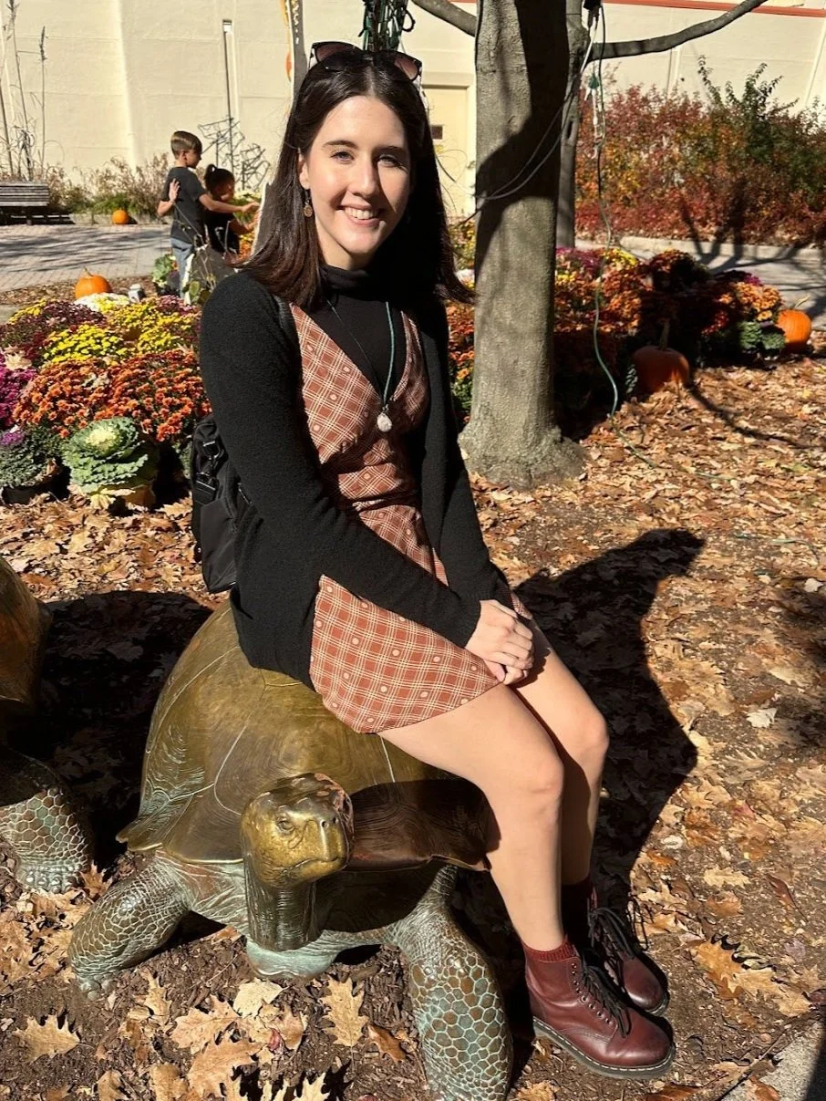 This is a picture of me, Ally Goetz. I have short brown hair and I am 22 years old. I am at the Toledo Zoo in the fall. I am sitting on a brass tortoise statue.
