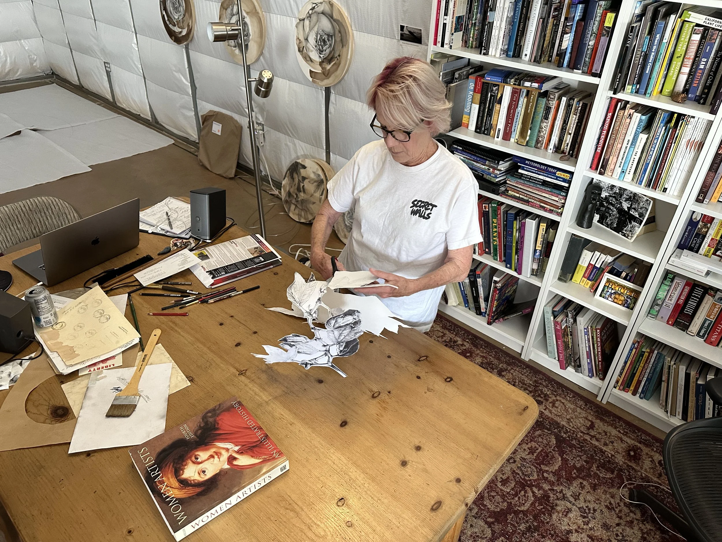 An older woman with blonde hair and glasses, wearing a white t-shirt with black text, working at a wooden table with art supplies and torn paper sketches, in a room with a bookshelf full of books.