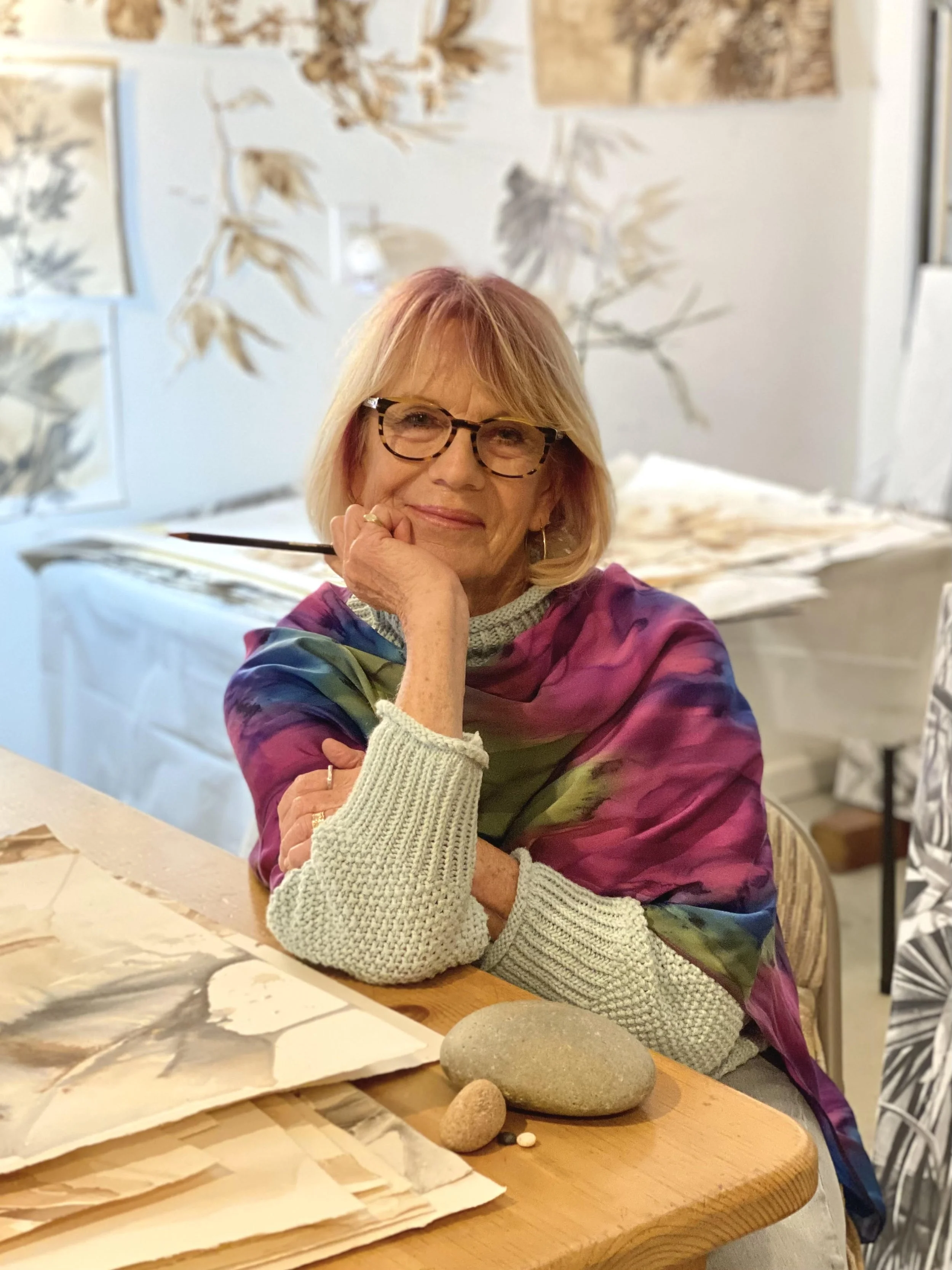 A woman with blonde hair and glasses manifests a colorful silk scarf draped over her shoulders. She sits at a wooden table, resting her face on her left hand, with a contemplative and gentle smile. The table has some papers, stones, and small objects. Behind her, a background with leaf and abstract art is partially visible.