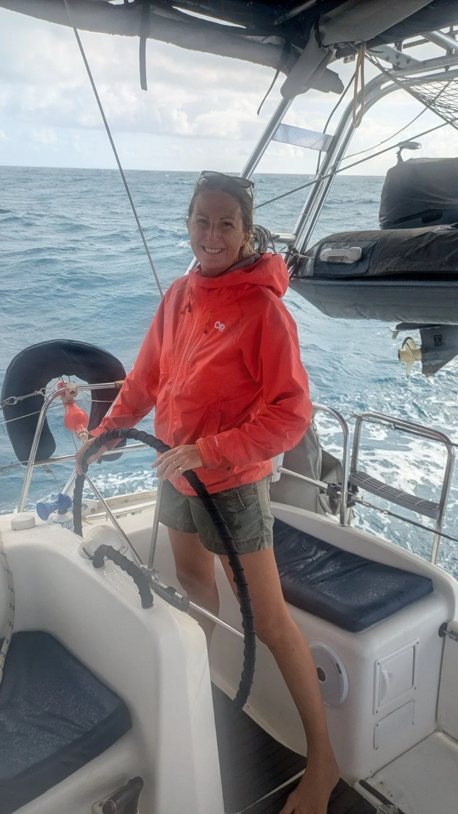 A woman smiling and steering a boat in the ocean during daytime, wearing a red jacket and shorts.