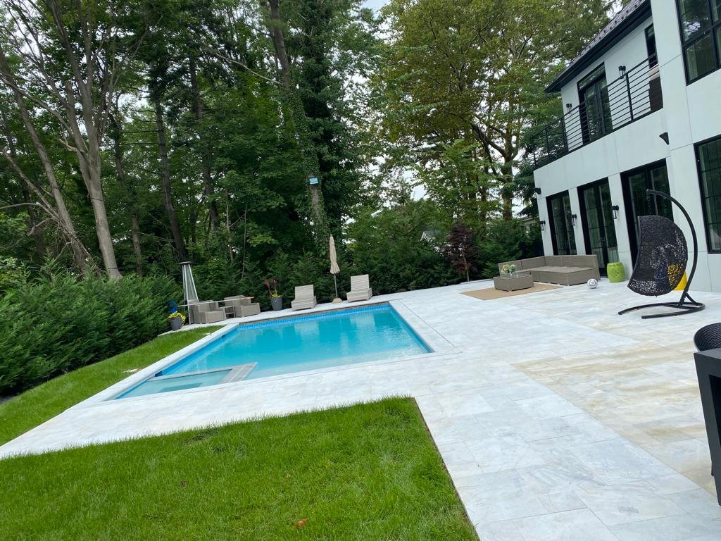 Modern backyard with a swimming pool, surrounded by a white patio, outdoor furniture, lounge chairs, and a tree line in the background.